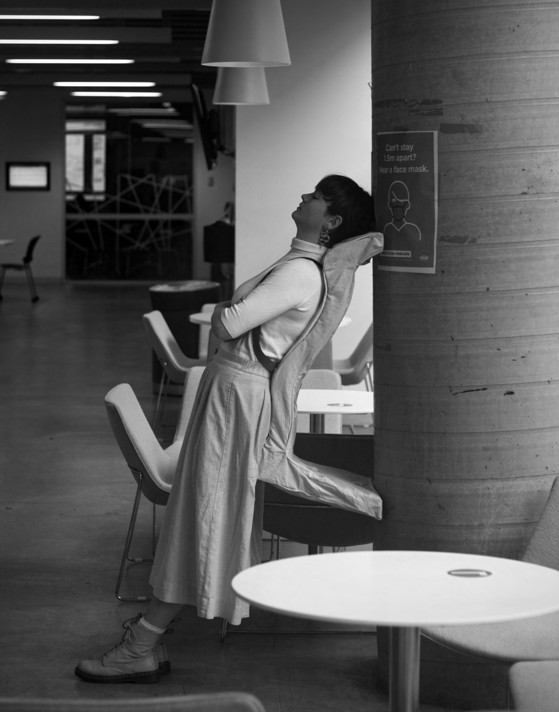 A woman with short hair and earrings leaning back in a chair with a neck pillow, wearing a light-colored turtleneck and baggy pants, in a modern cafe or waiting area, with a face mask safety sign on a pillar nearby.