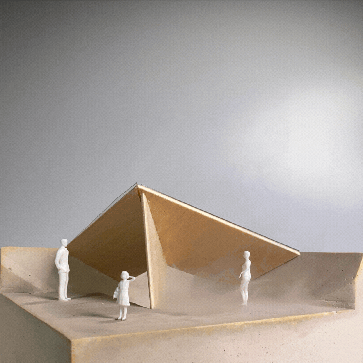 Miniature architectural model with a large, angled wooden roof supported by a single vertical wall, and four small white figurines of people around it, on a flat surface against a plain background.