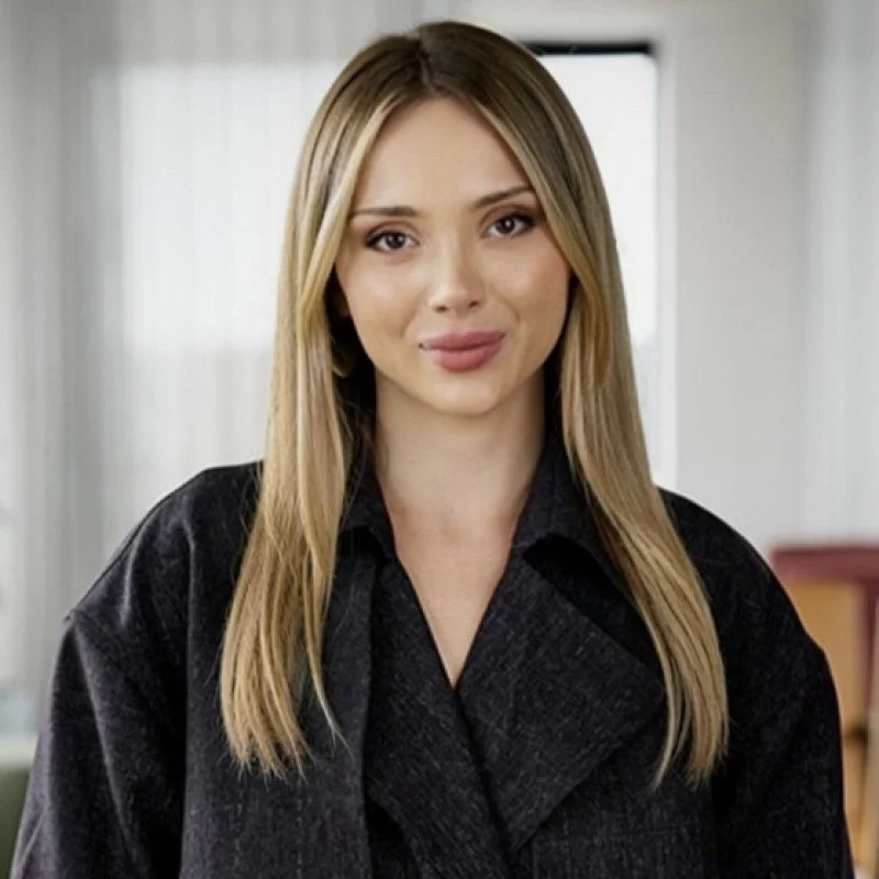 Portrait of a young woman with long blonde hair, wearing a dark jacket, smiling softly, in an indoor setting.