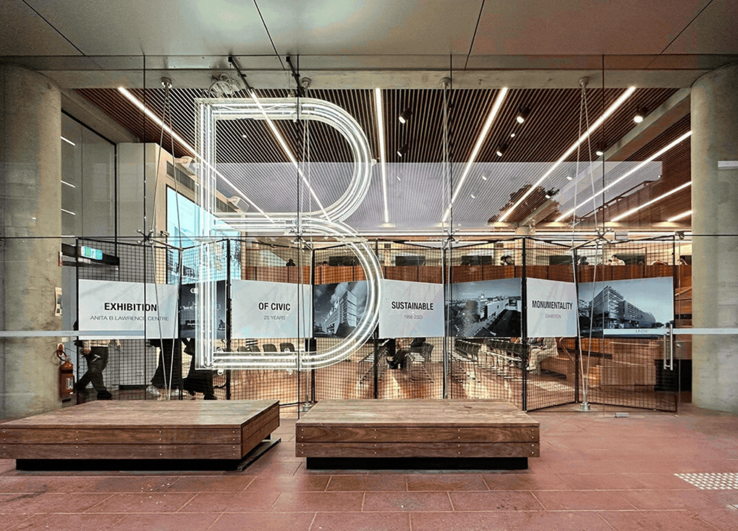 Interior view of a modern building with a large illuminated '3' sign, glass panels with text about an exhibition, and people inside. The environment features wood and concrete design elements.