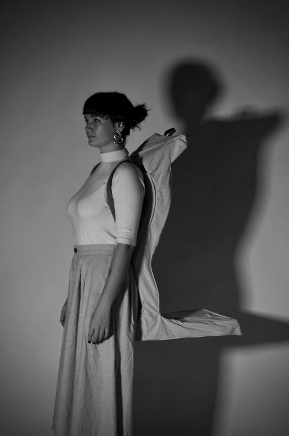 A woman with short hair and large earrings stands in front of a plain background, with her shadow cast on the wall behind her. She wears a long-sleeved turtleneck and high-waisted trousers, with a backpack on her shoulders.