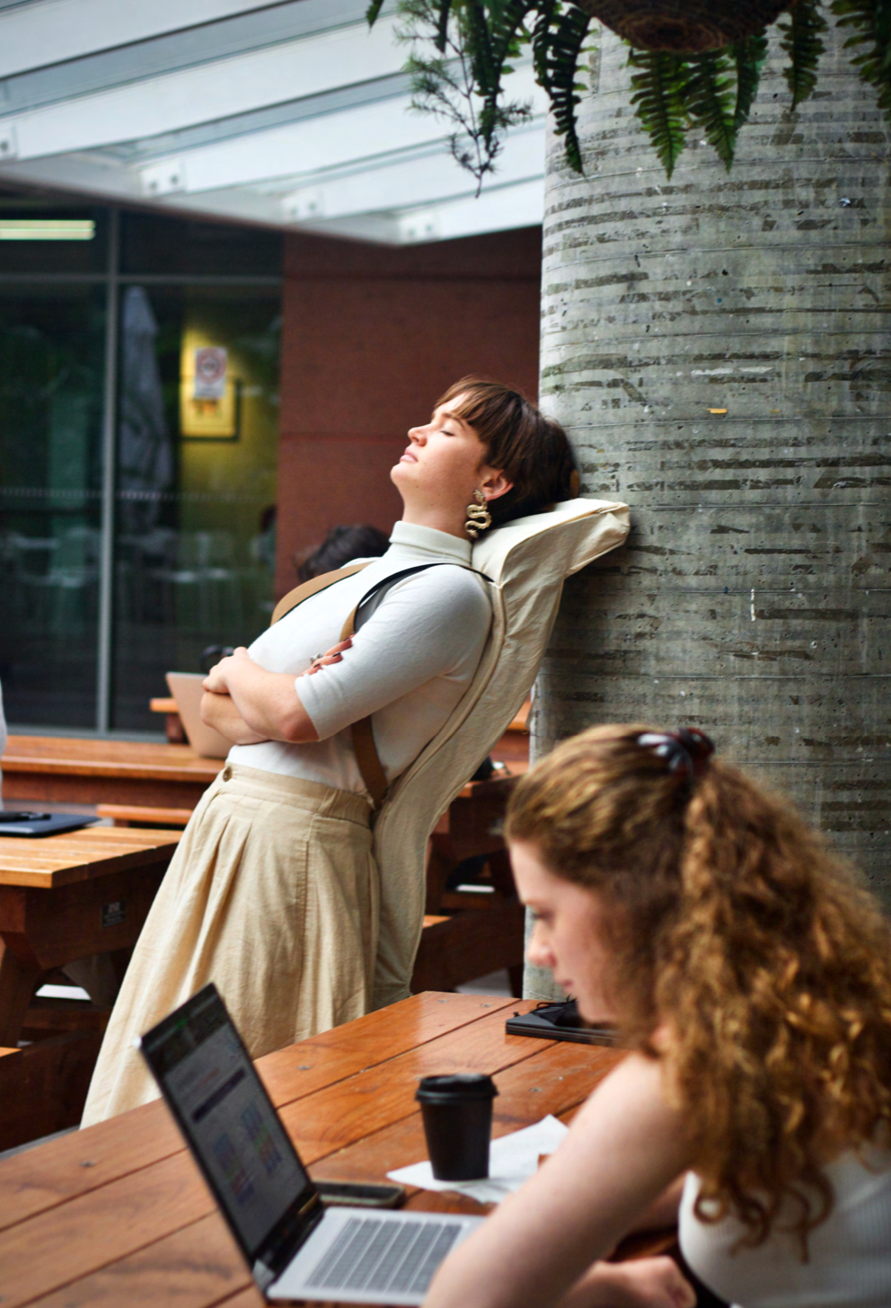 A woman leans back in a chair with her eyes closed, resting against a large, textured concrete pillar in a cafe or public space. Another woman with curly red hair is working on a laptop at a wooden table in the foreground.