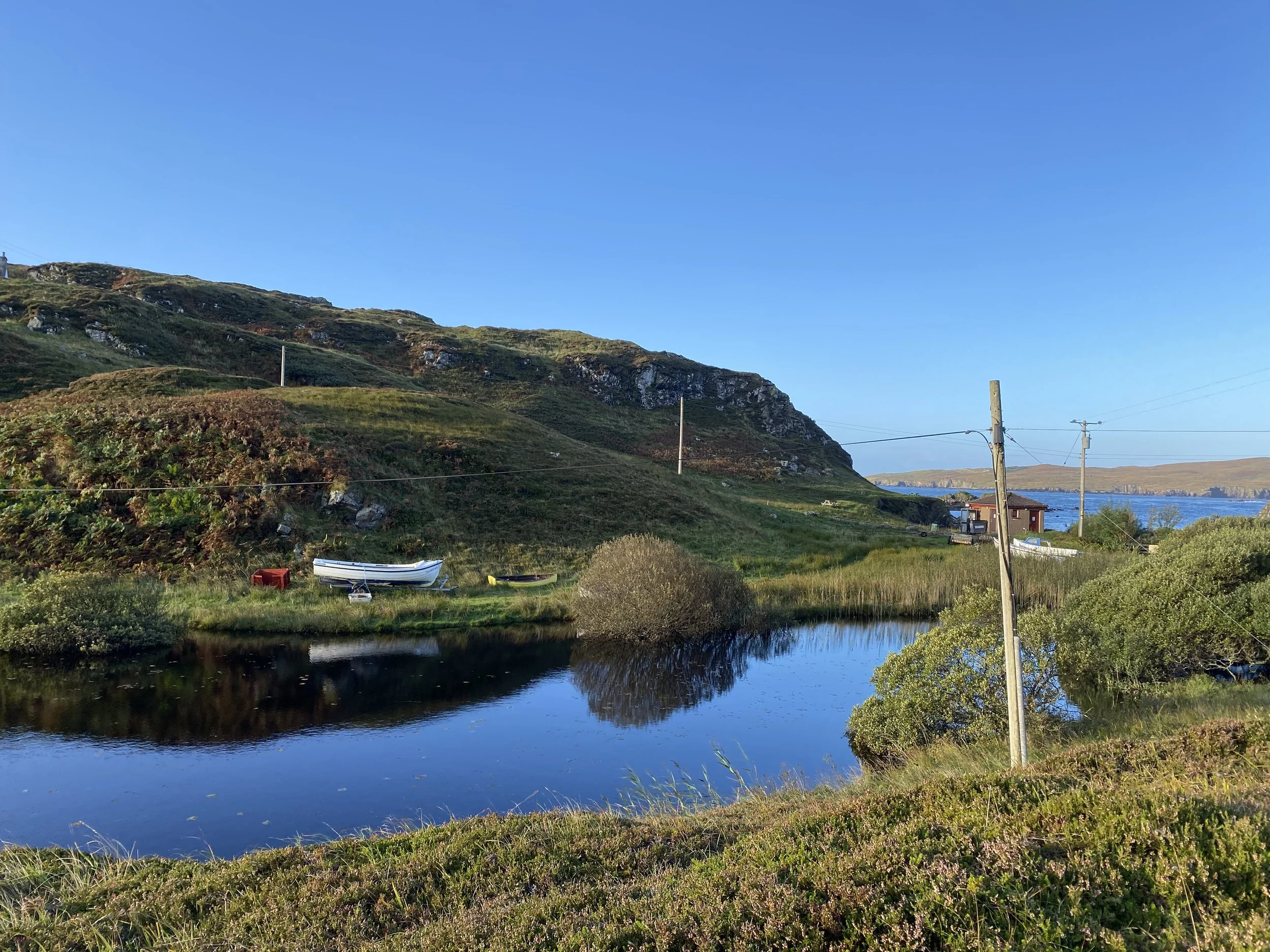 A scenic landscape featuring a calm river reflecting the blue sky, green hillside with bushes, and some boats along the riverbank. There are utility poles and a small house near the water with hills in the background.