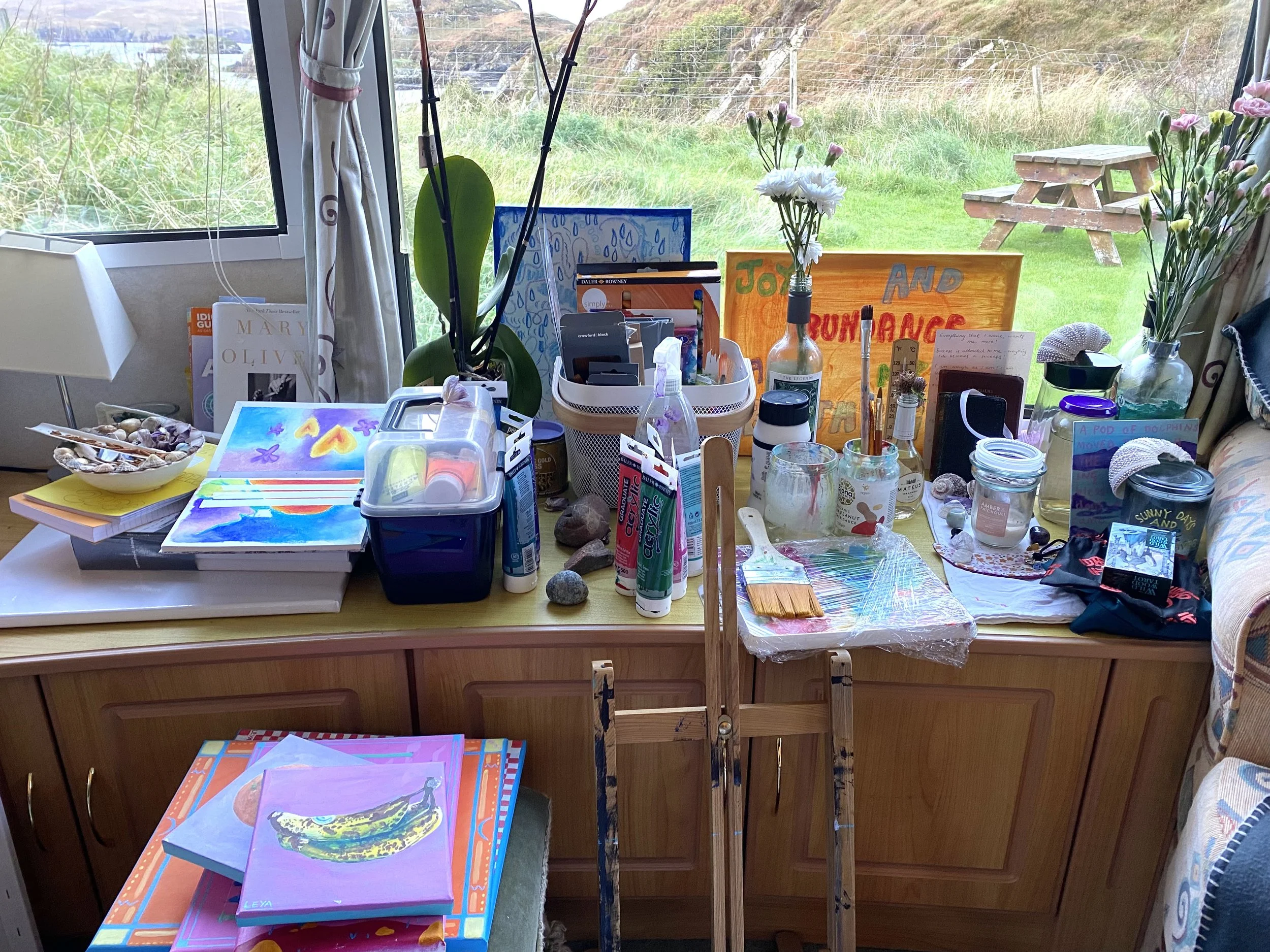 Indoor desk filled with art supplies, paintings, books, and decorative items, with a large window overlooking a grassy outdoor area with a picnic table and a wire fence.