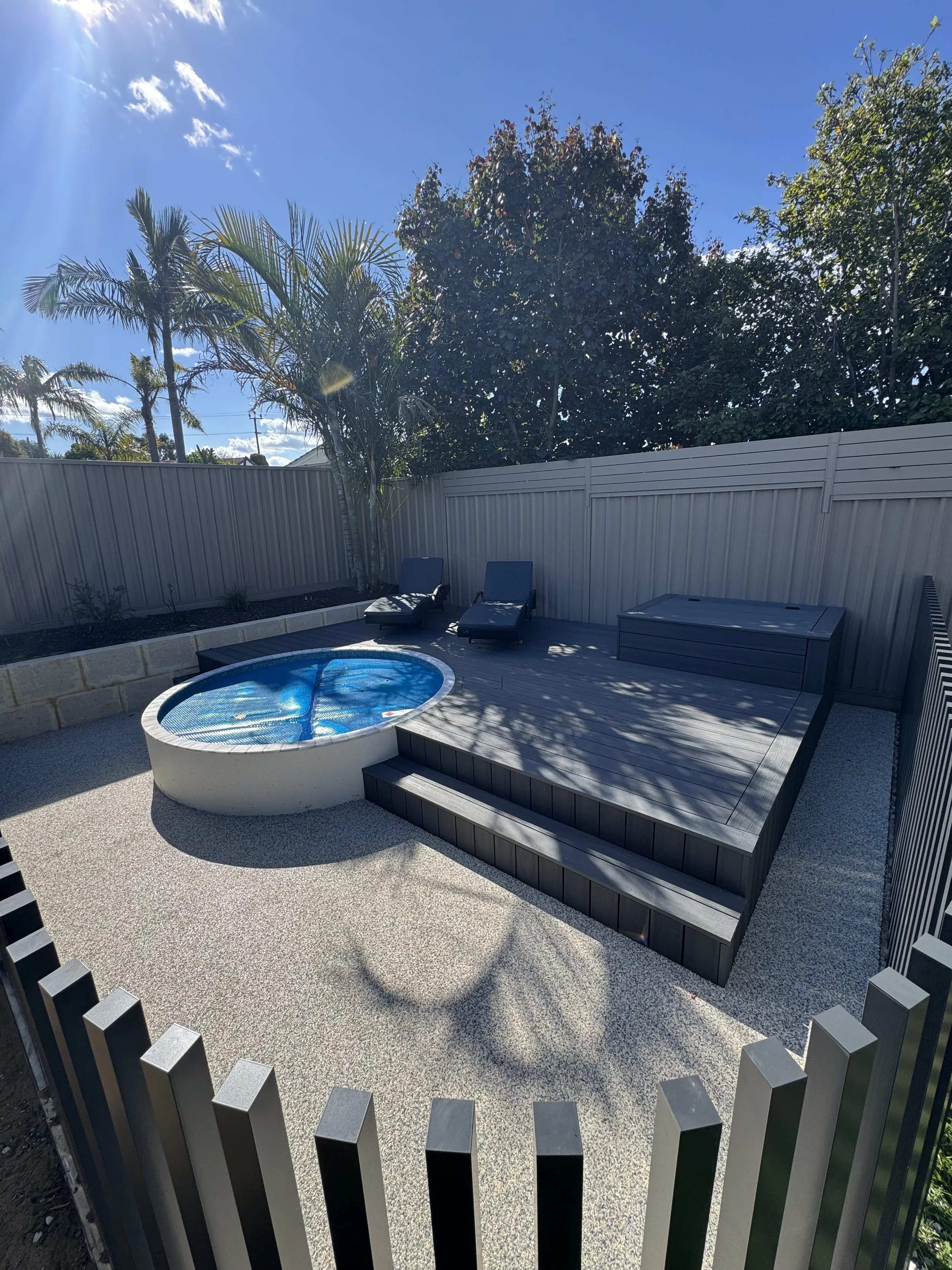 A backyard patio with a small round pool, two lounge chairs, a wooden platform, a storage box, a tan fence, and tall trees including palm trees against a blue sky with some clouds.