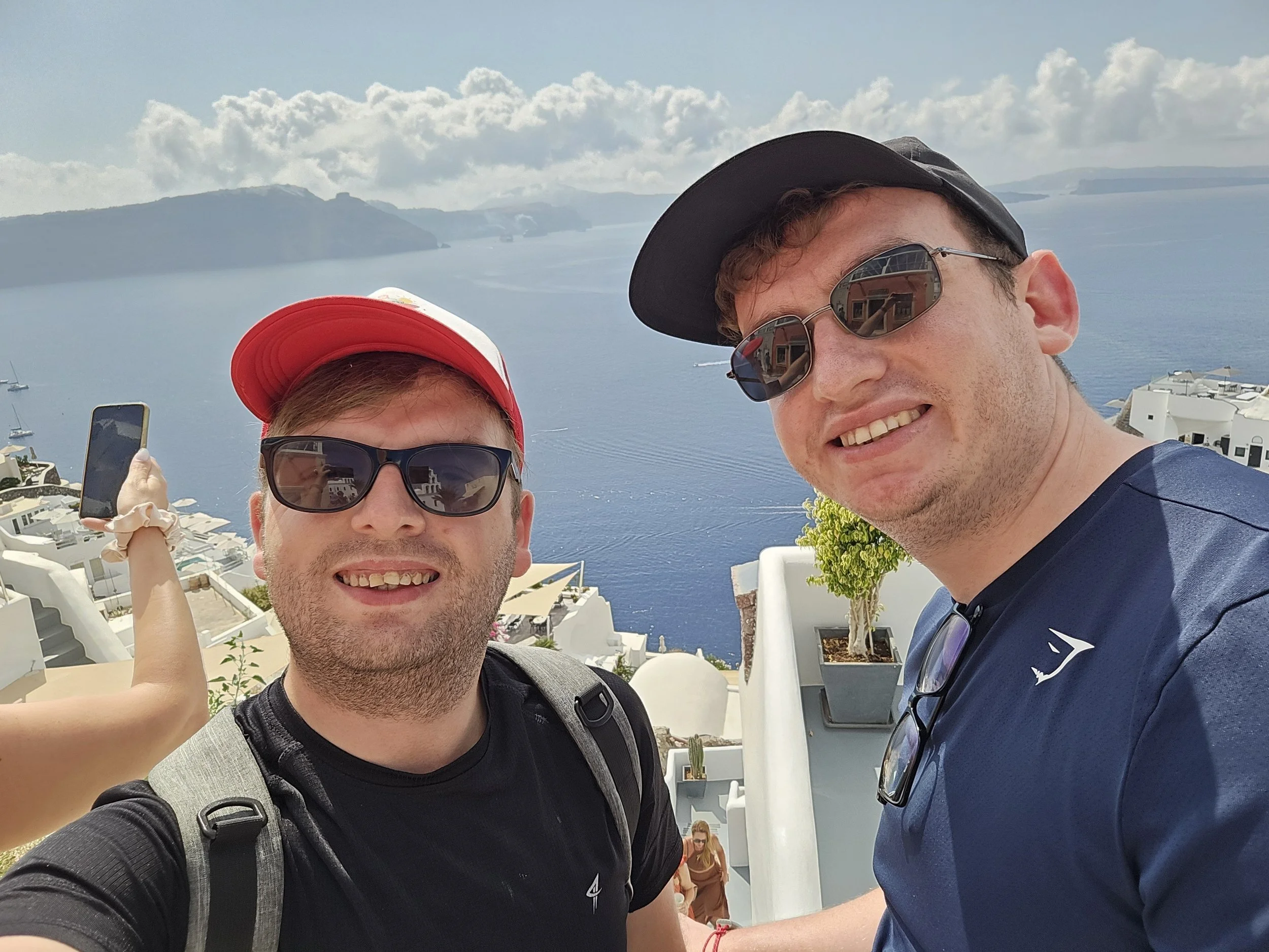 Two young men wearing sunglasses and hats taking a selfie with a scenic water view and white buildings in the background.