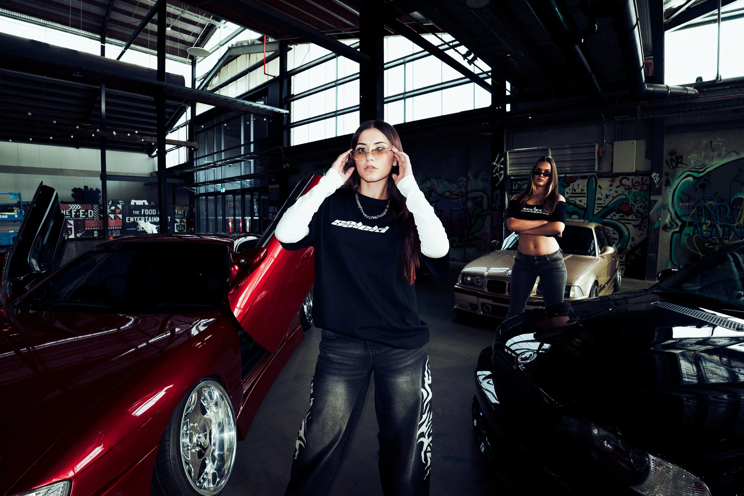 Two women are posing near sports cars inside a garage with graffiti walls and large windows. The woman in the foreground is adjusting her sunglasses and wearing a black and white shirt, while the woman in the background is standing with her arms crossed.