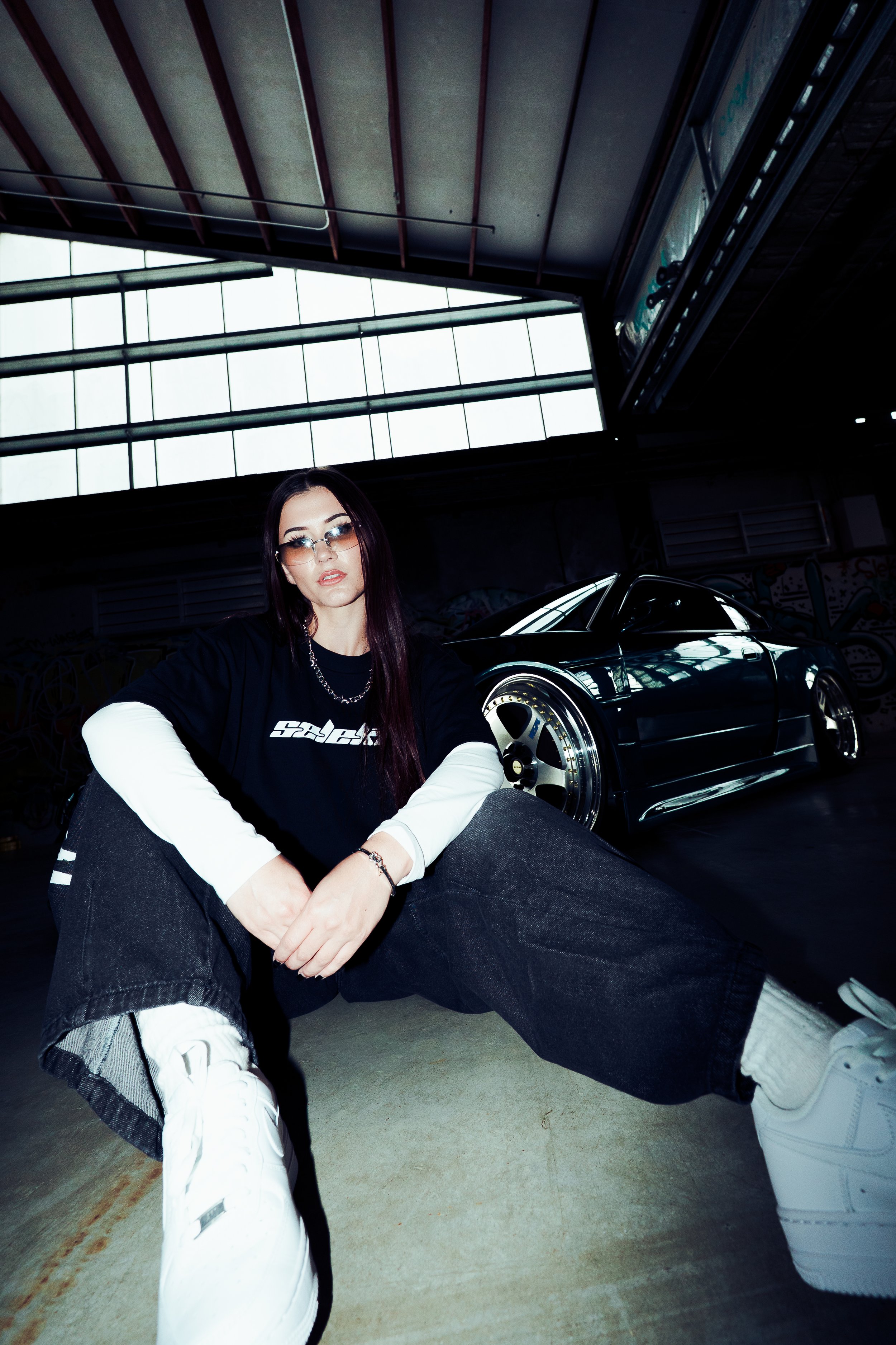 A young woman with long purple hair and sunglasses is sitting on the ground in an industrial-style parking garage. She is wearing a black long-sleeve shirt with white sleeves underneath, dark jeans, and white sneakers. Behind her, there is a black sports car and large windows that let in natural light.