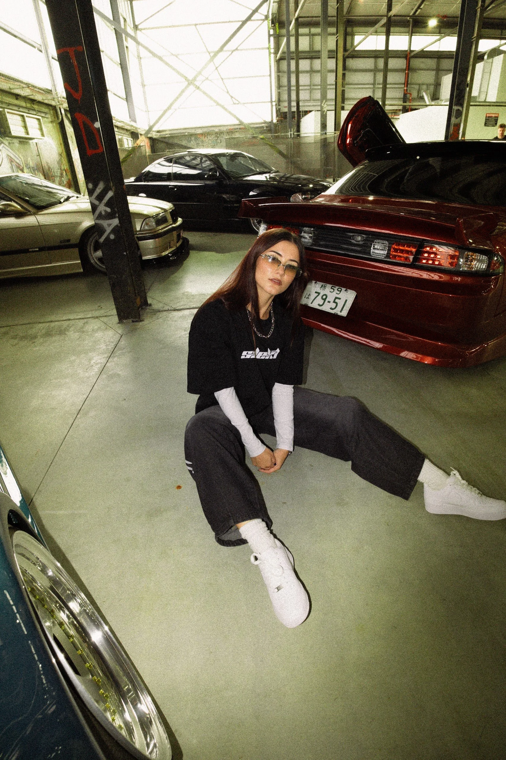 A young woman with long dark hair, wearing glasses, a black t-shirt, black pants, and white sneakers, is sitting on the floor of a parking garage next to a red sports car with the trunk open. She is looking slightly to the side with a neutral expression.