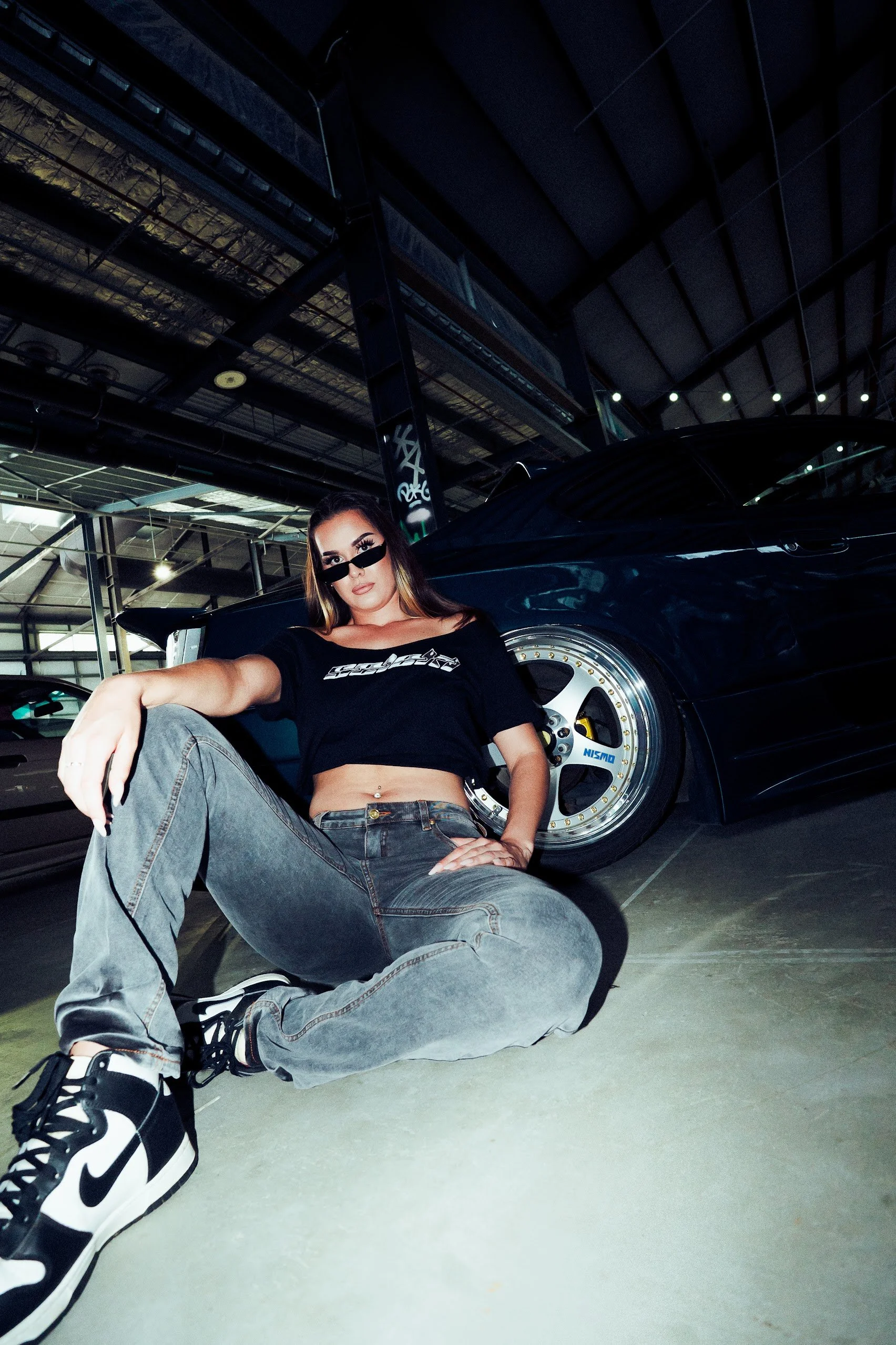 A woman wearing sunglasses, a black cropped T-shirt, and baggy gray jeans, sitting on the floor of a parking garage next to a black sports car with aftermarket wheels.