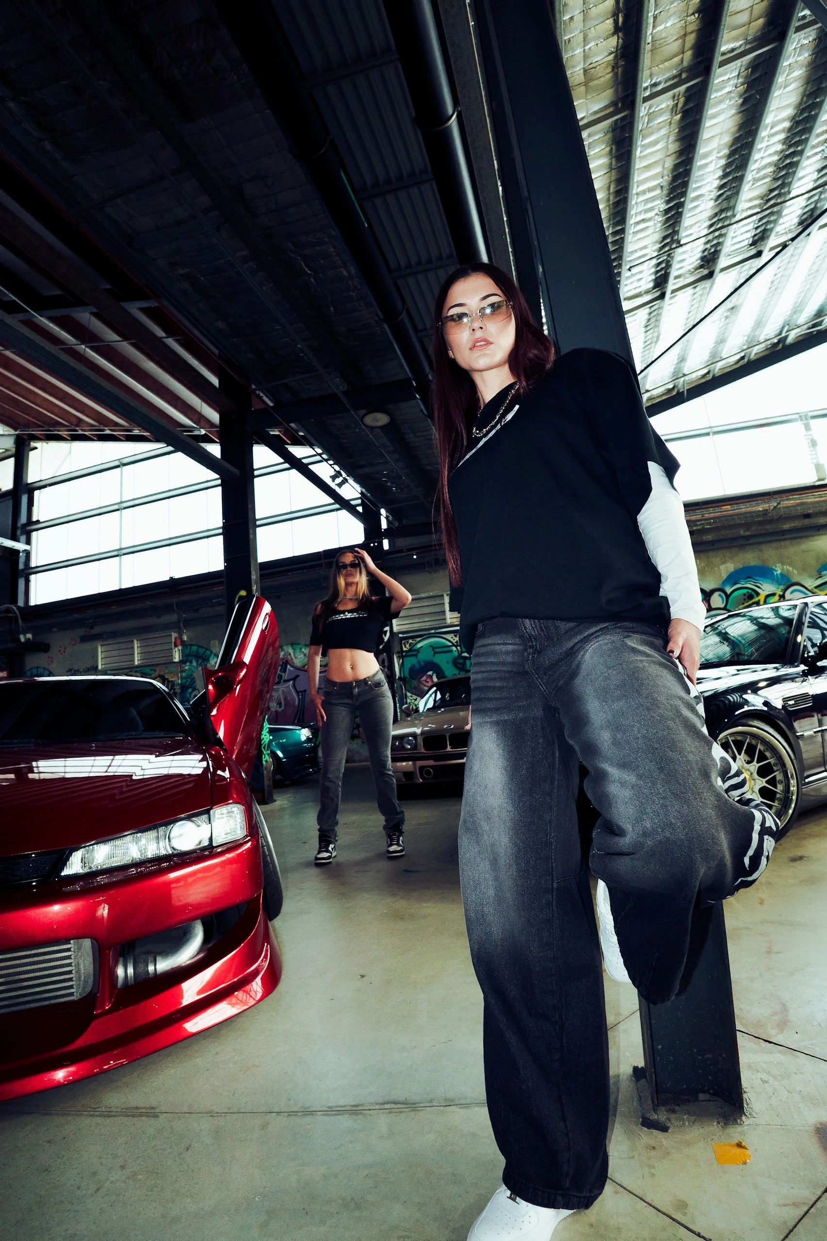 Two women posing near a red sports car in an indoor parking garage with graffiti on the walls and large windows above.