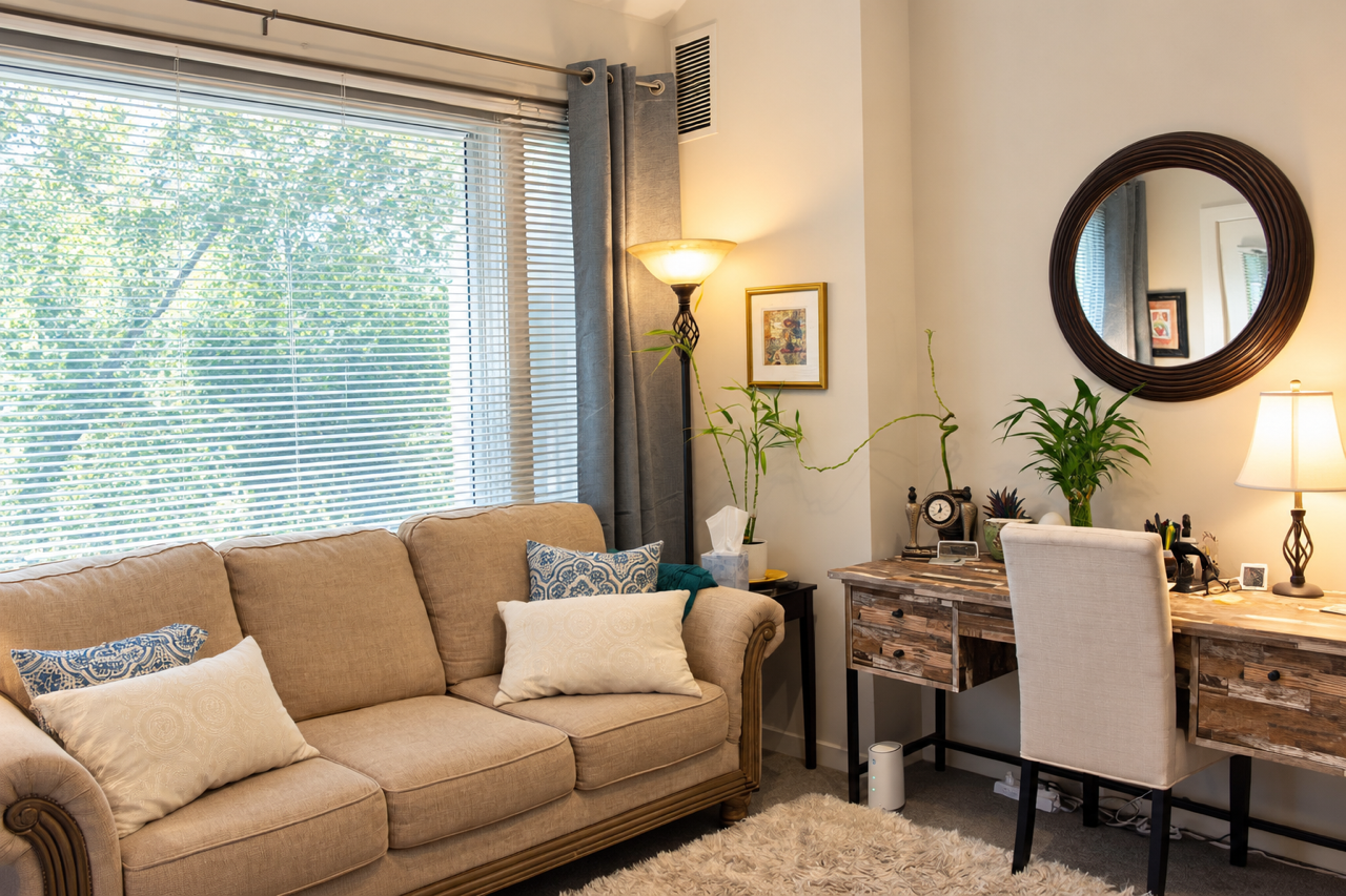 Living room with beige sofa, decorative pillows, large window with blinds, desk with plants, mirror, lamp, and framed artwork.
