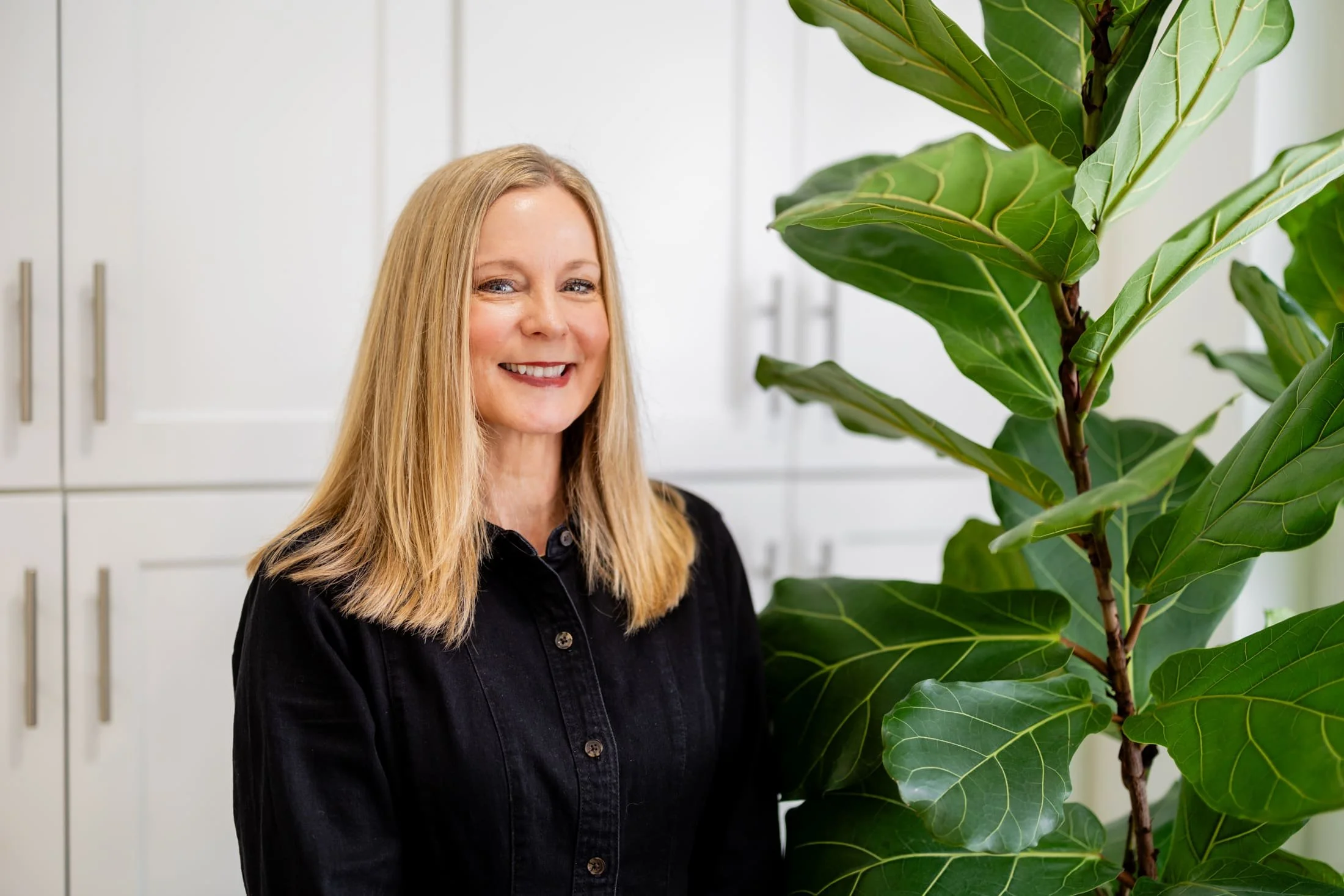 A woman with blonde hair and a black shirt smiling next to a large green leafy plant in a modern indoor setting.