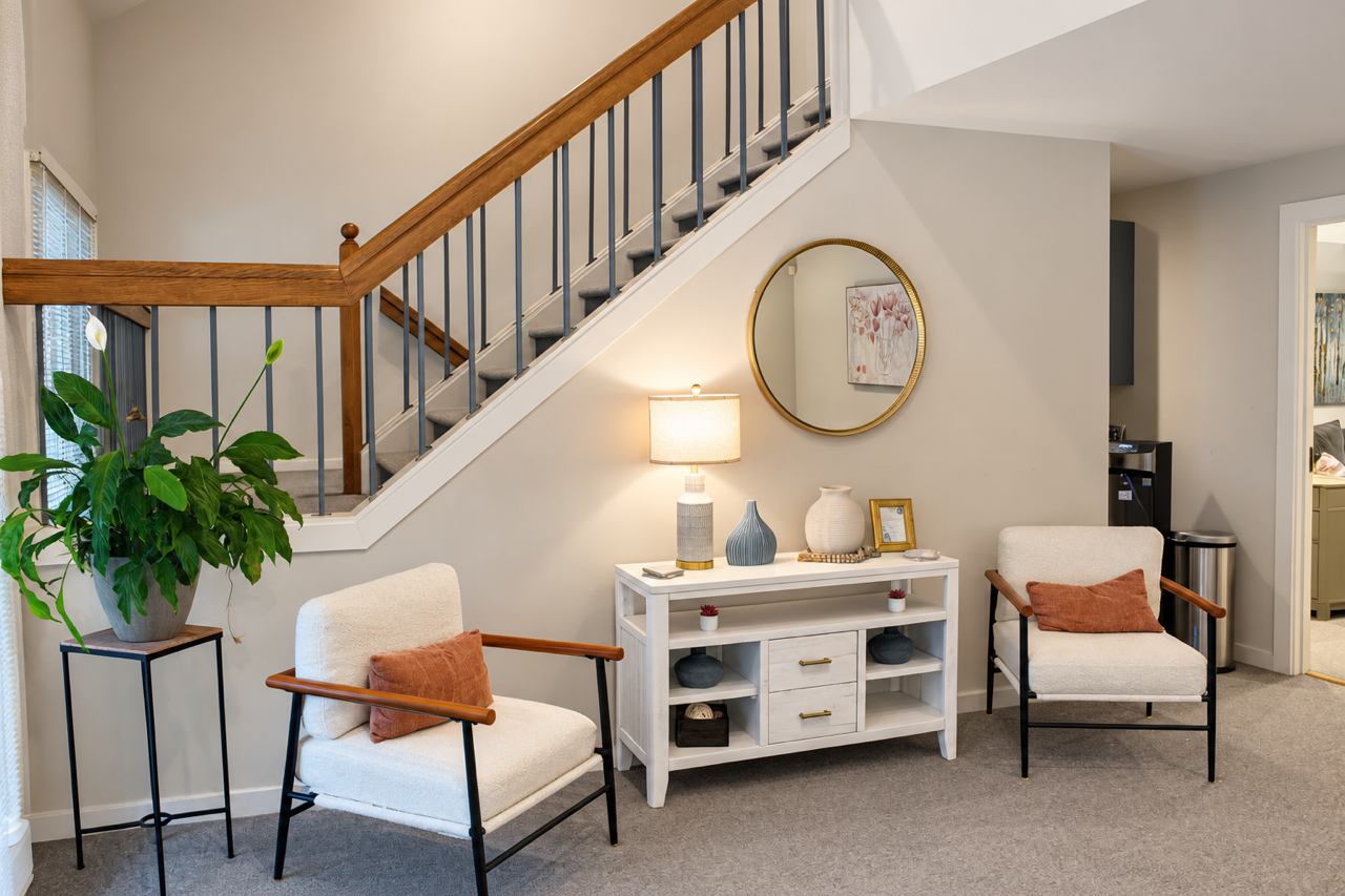 Living room area with a white console table, two beige chairs with wooden armrests, a large potted plant on a black stand, a lamp, decorative vases, framed photo, and a round mirror on the wall under a staircase with wooden railing.