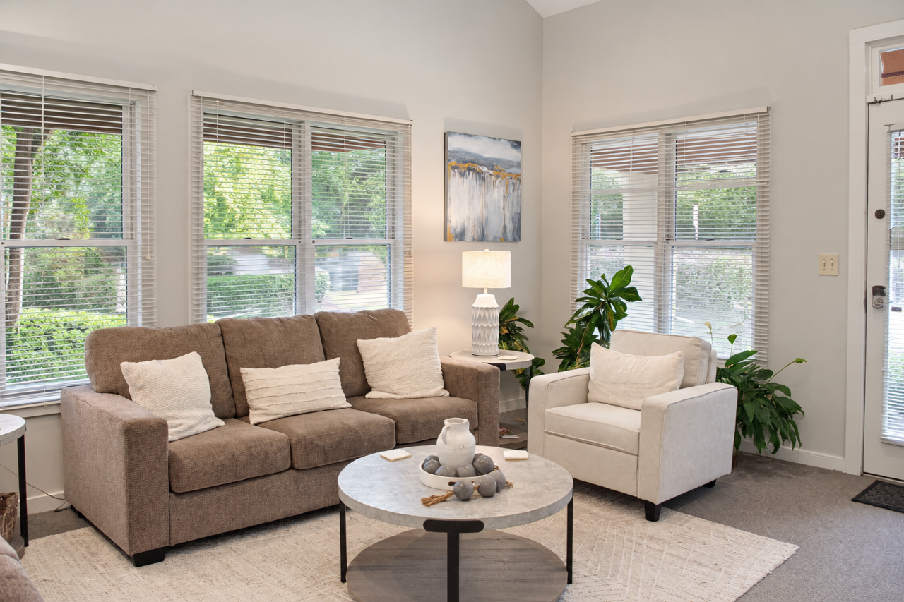 Living room with beige sofa, white armchair, coffee table, lamp, and green plants by windows and door