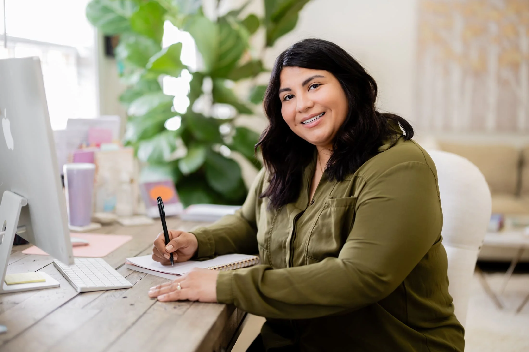 A woman with long black hair, wearing an olive green shirt, sitting at a desk with a computer, notepad, and pen, smiling at the camera in a bright, plant-filled office.