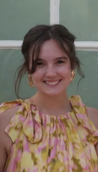 A young woman with brown hair styled in loose waves, wearing gold earrings and a colorful yellow and pink floral dress, smiling in front of a green wall with white trim.