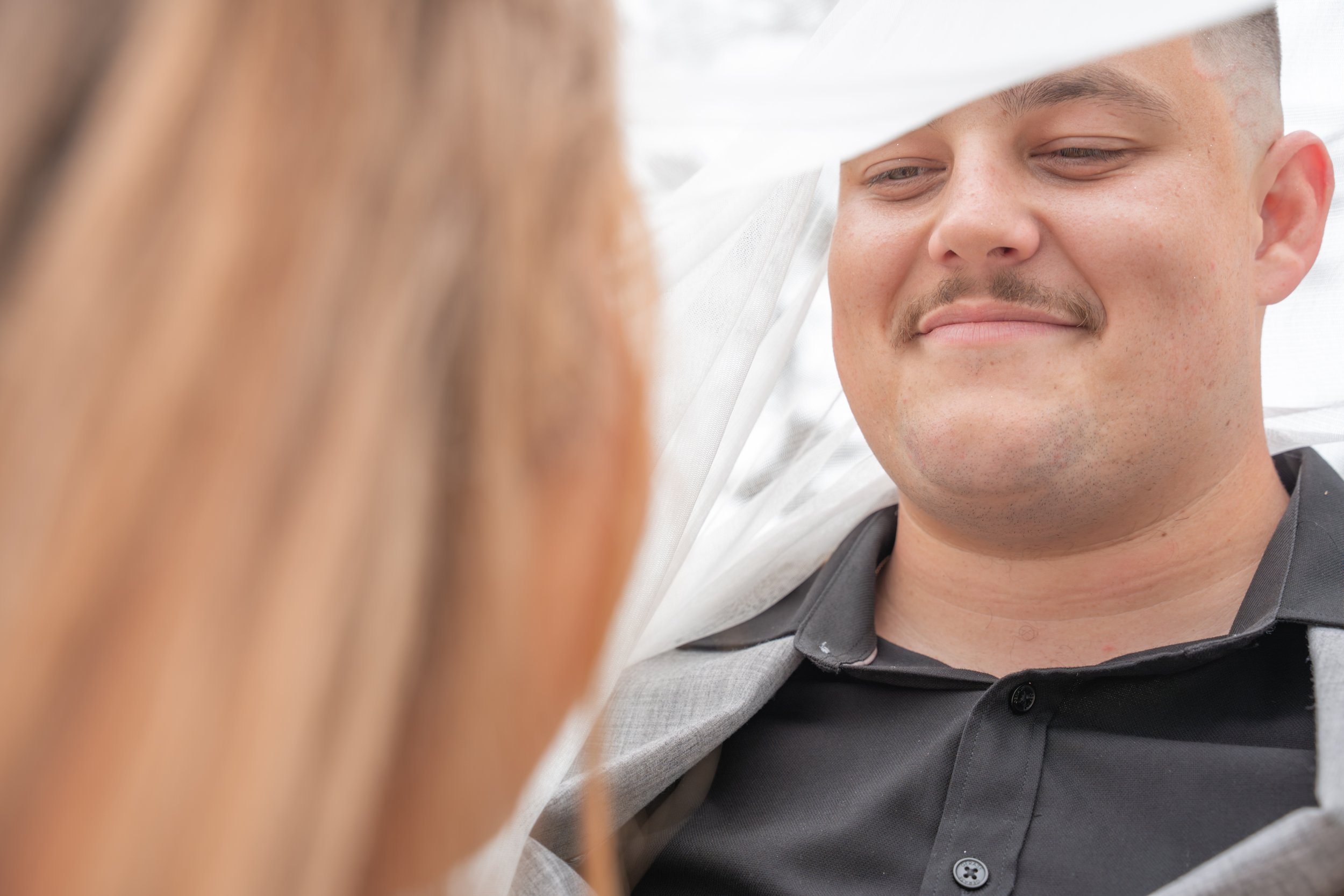 Close-up of a smiling man in a gray suit smiling under wife's veil, with a woman with brown hair partially visible in the foreground.