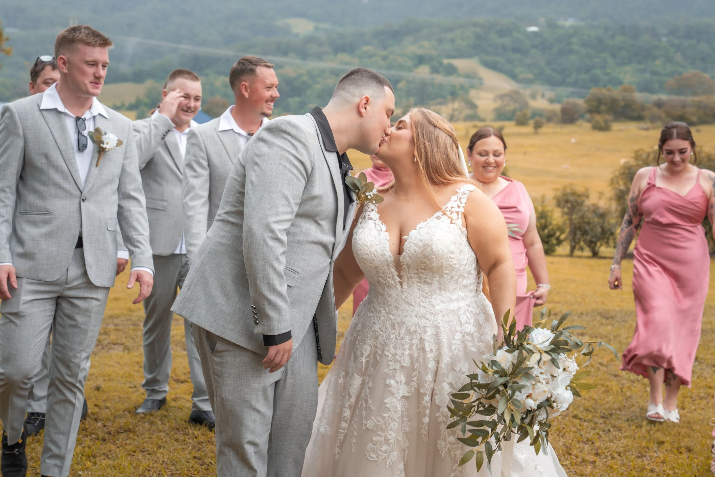 A newly married couple shares a kiss at their outdoor wedding, surrounded by friends in pastel and light gray suits, with rolling green hills in the background.