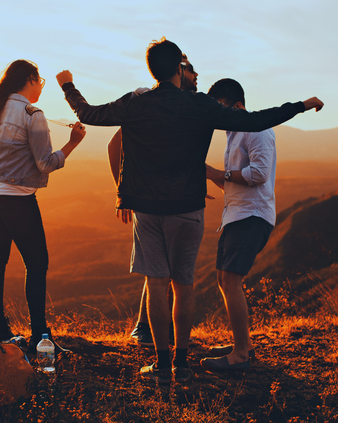 Gruppe von fünf jungen Menschen, die bei Sonnenuntergang im Freien stehen, einige schauen sich an, andere unterhalten sich fröhlich, auf einem Hügel mit einer Landschaft im Hintergrund.