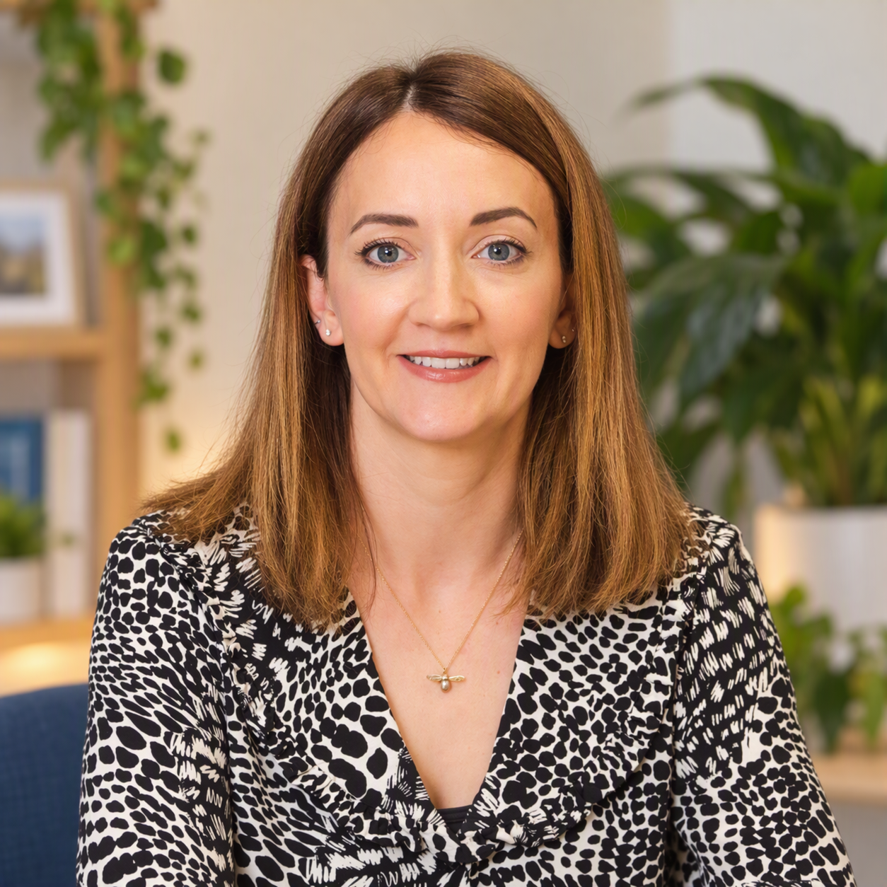 Portrait of a woman with shoulder-length brown hair, wearing a black and white patterned blouse, gold necklace, and earrings, sitting in a modern office with plants and shelves in the background.