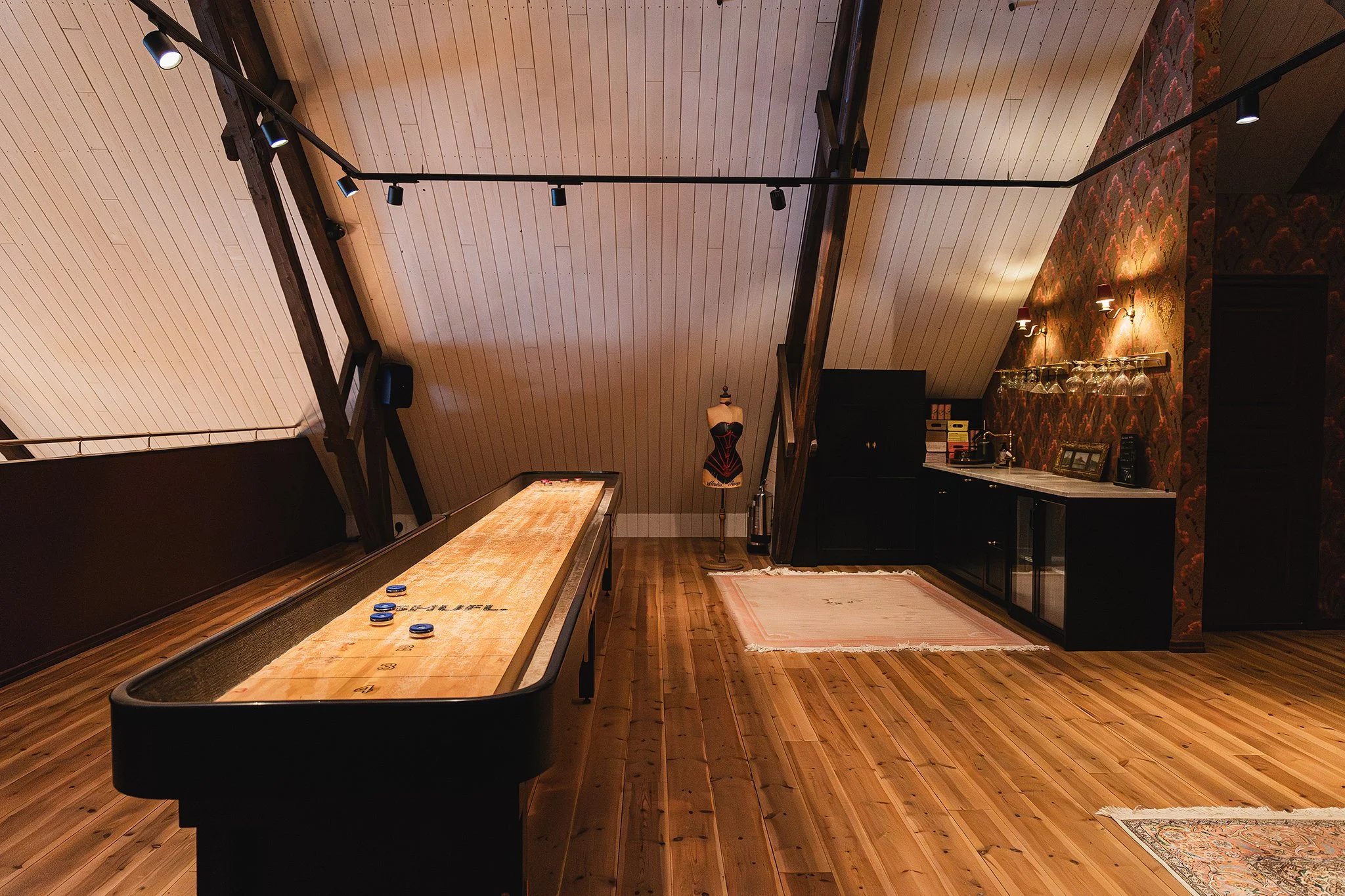Indoor game room with wood-paneled sloped ceiling, a shuffleboard table with game pieces, bar area with glasses hanging, decorative wallpaper with floral pattern, mannequin wearing black and red clothing, and vintage-style lamp.