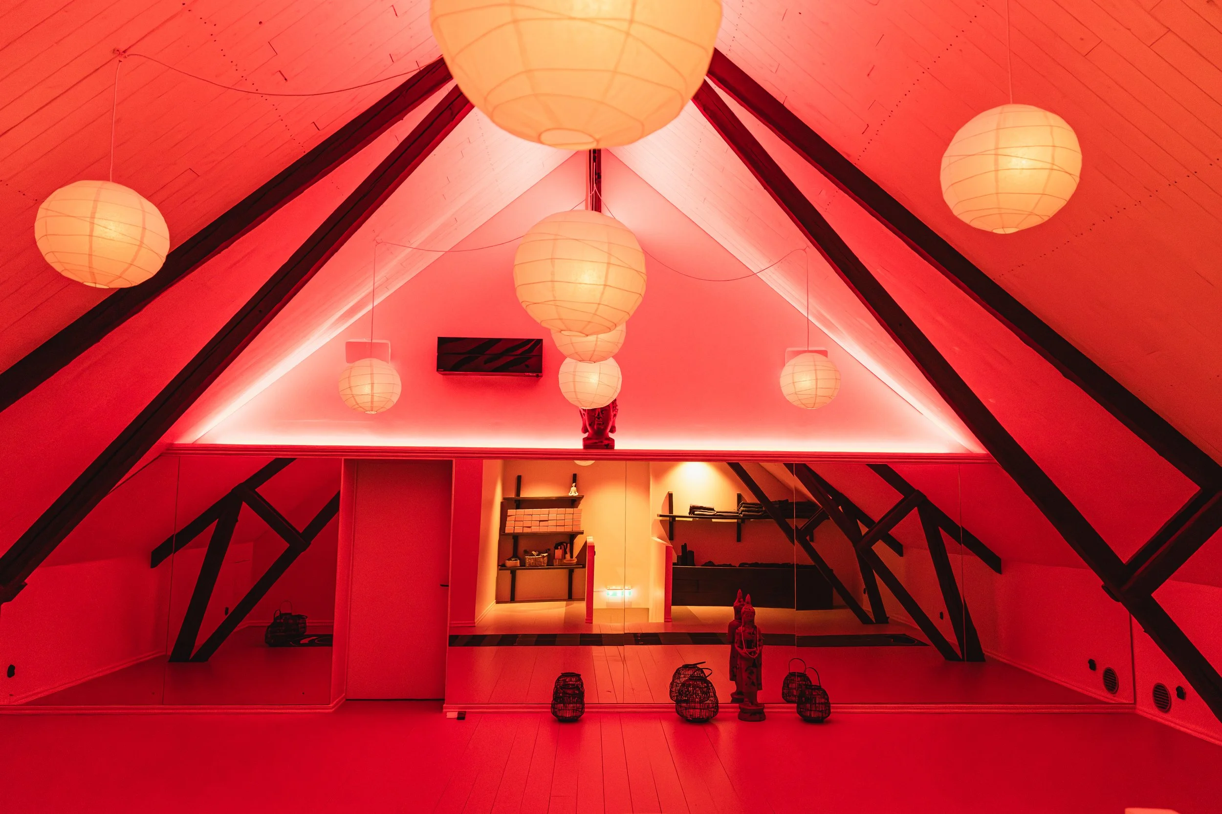 Interior view of a room with a vaulted ceiling painted red. Hanging paper lanterns are suspended from the ceiling. The room features black structural beams and a few decorative items, including lanterns and a small statue, with warm ambient lighting.