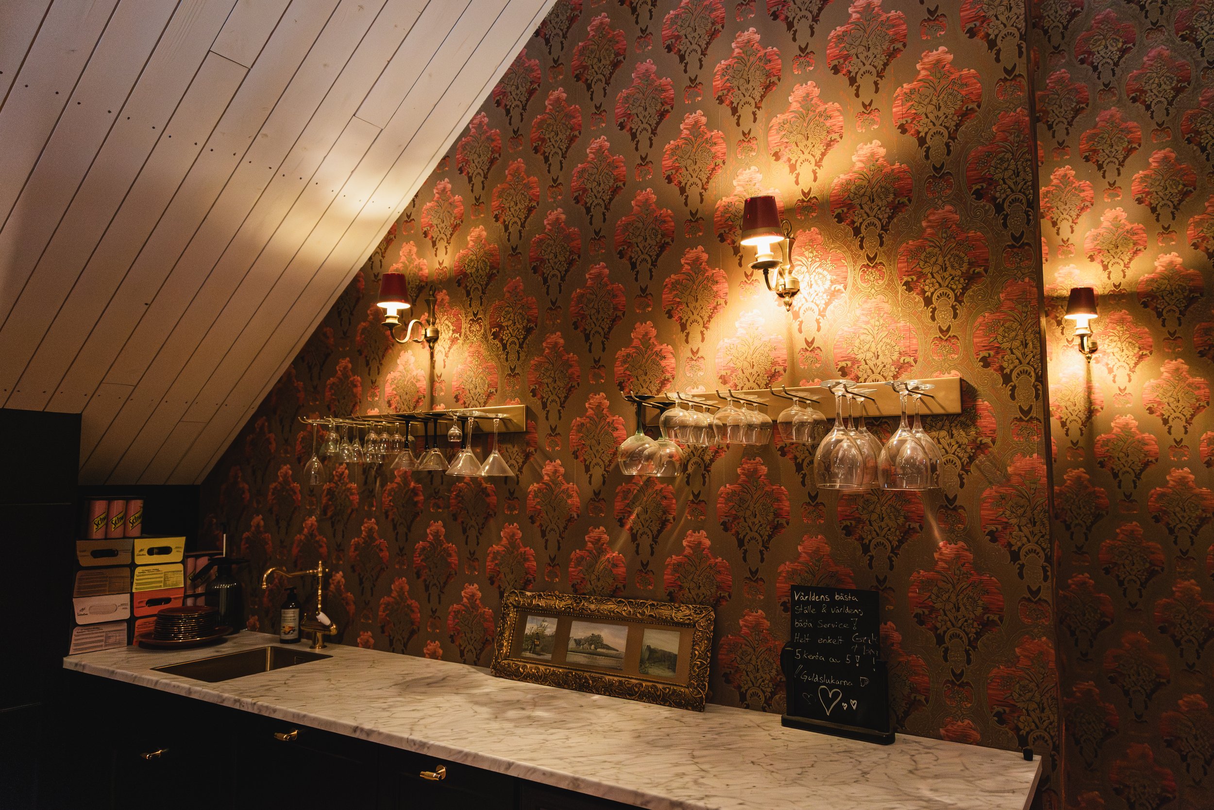 A bar area with a marble countertop, wine glasses hanging upside down, vintage-style wall lamps, and ornate wallpaper with a red and gold pattern.