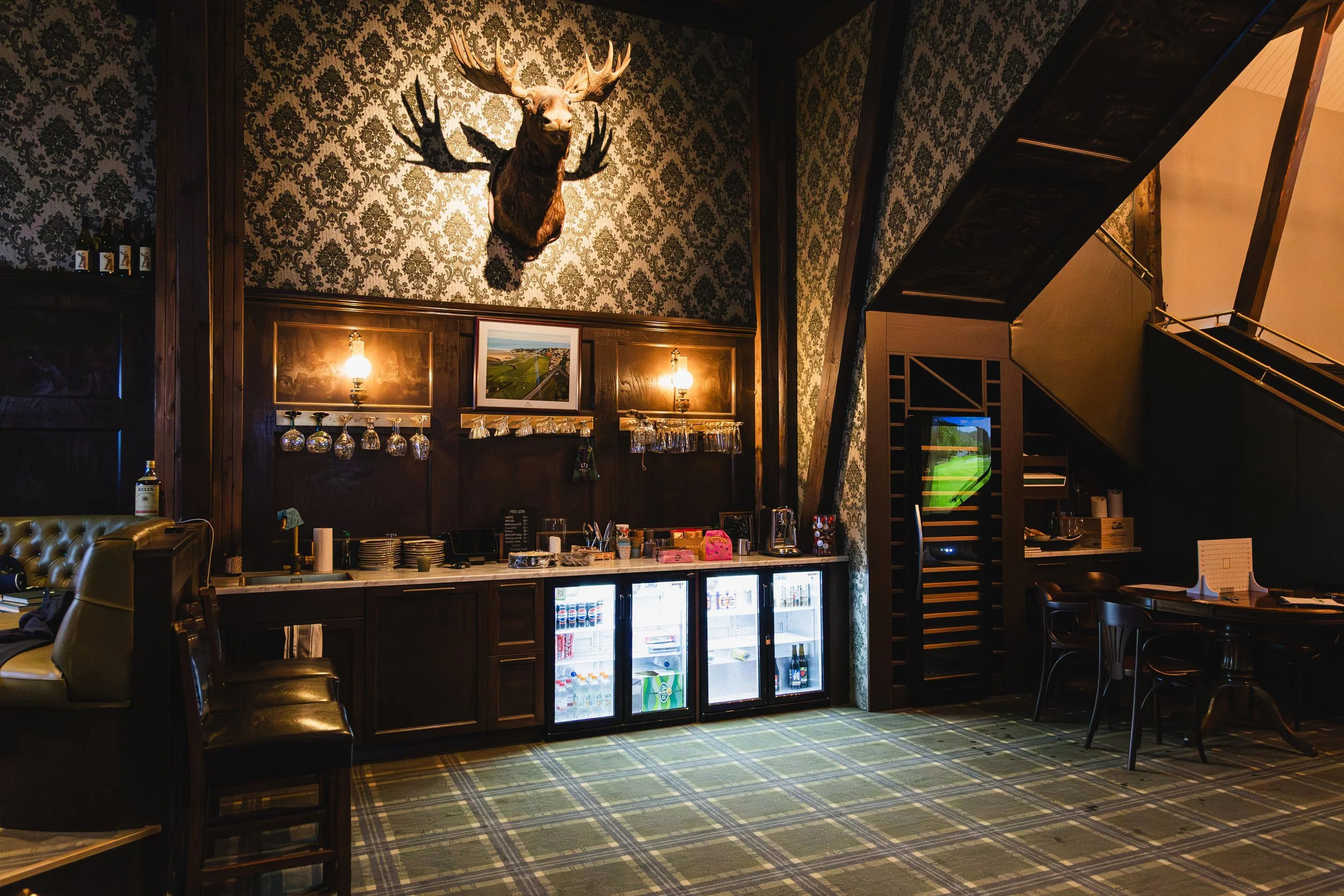 Interior of a bar or restaurant with a mounted deer head on the wall, dark wood furniture, a patterned carpet, and a glass-front refrigerator stocked with drinks.