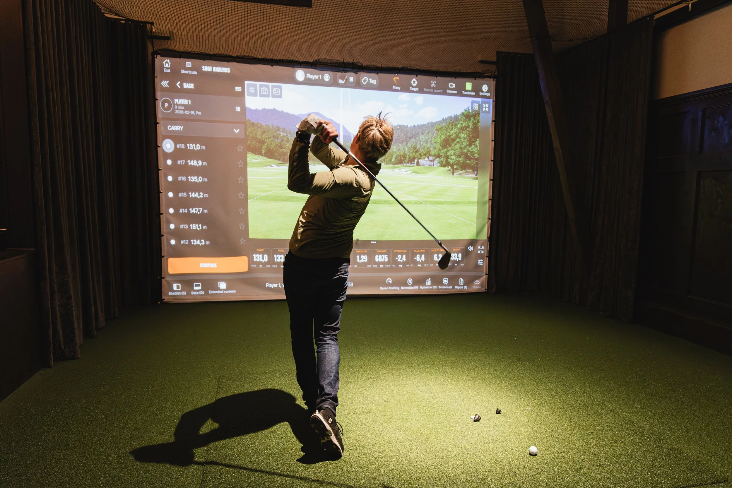 A person playing indoor golf simulation, swinging a golf club, in front of a large screen displaying a virtual golf course.