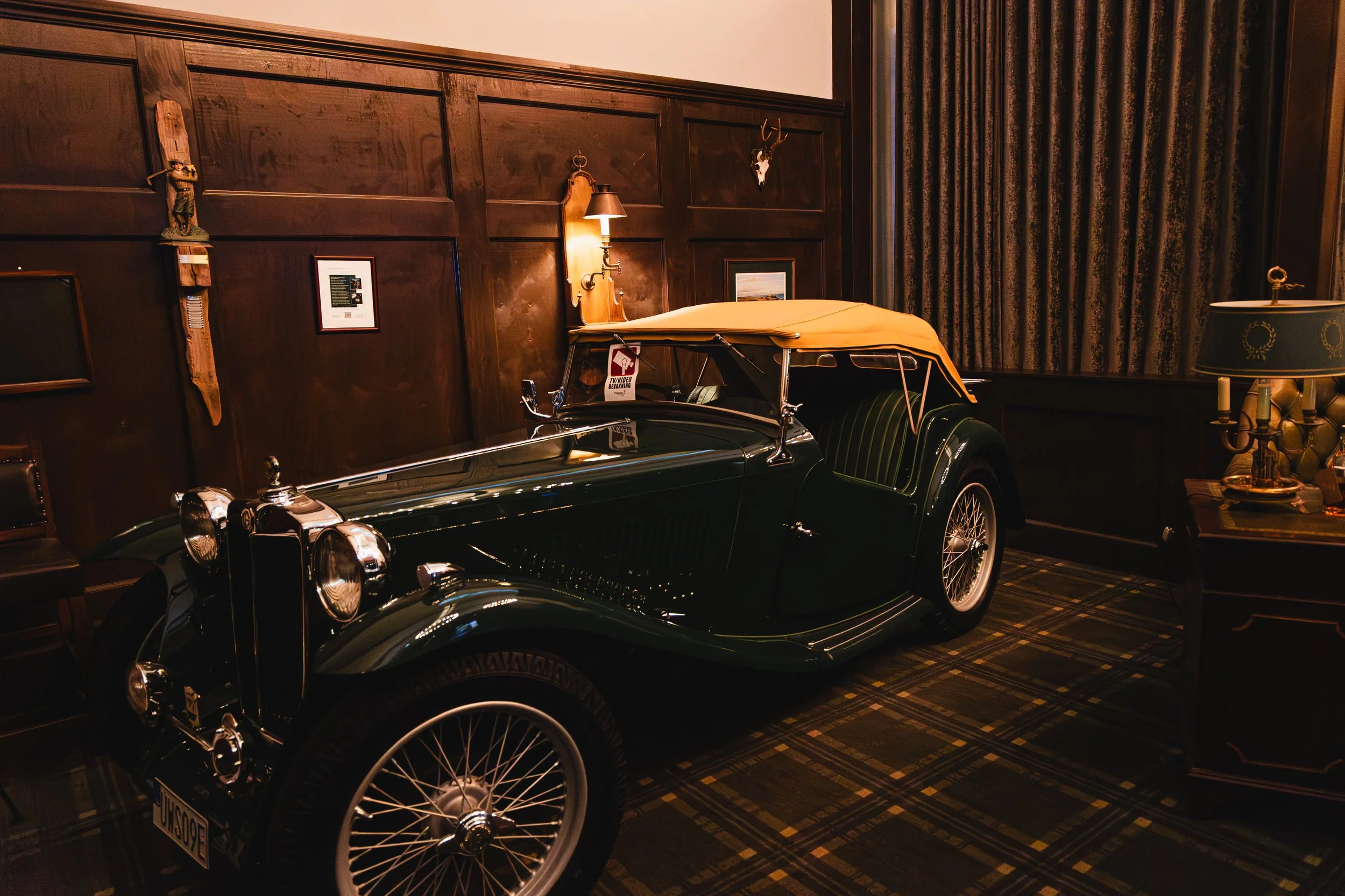 A vintage black convertible car with a yellow cover on the passenger side, parked indoors next to a dark wood-paneled wall. Decor includes a small statue, framed pictures, a wall lamp, and a mounted deer skull with antlers. There is also an ornate lamp on a table to the right.