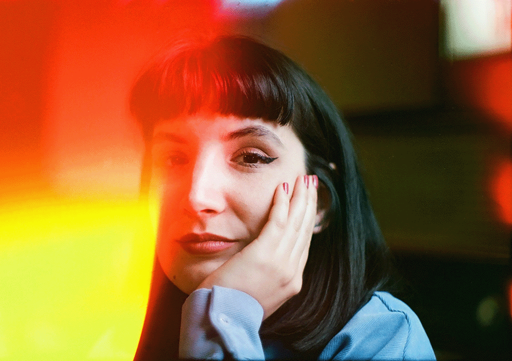 Close-up of a young woman with dark hair and makeup, resting her face on her hand in a colorful, blurred background.