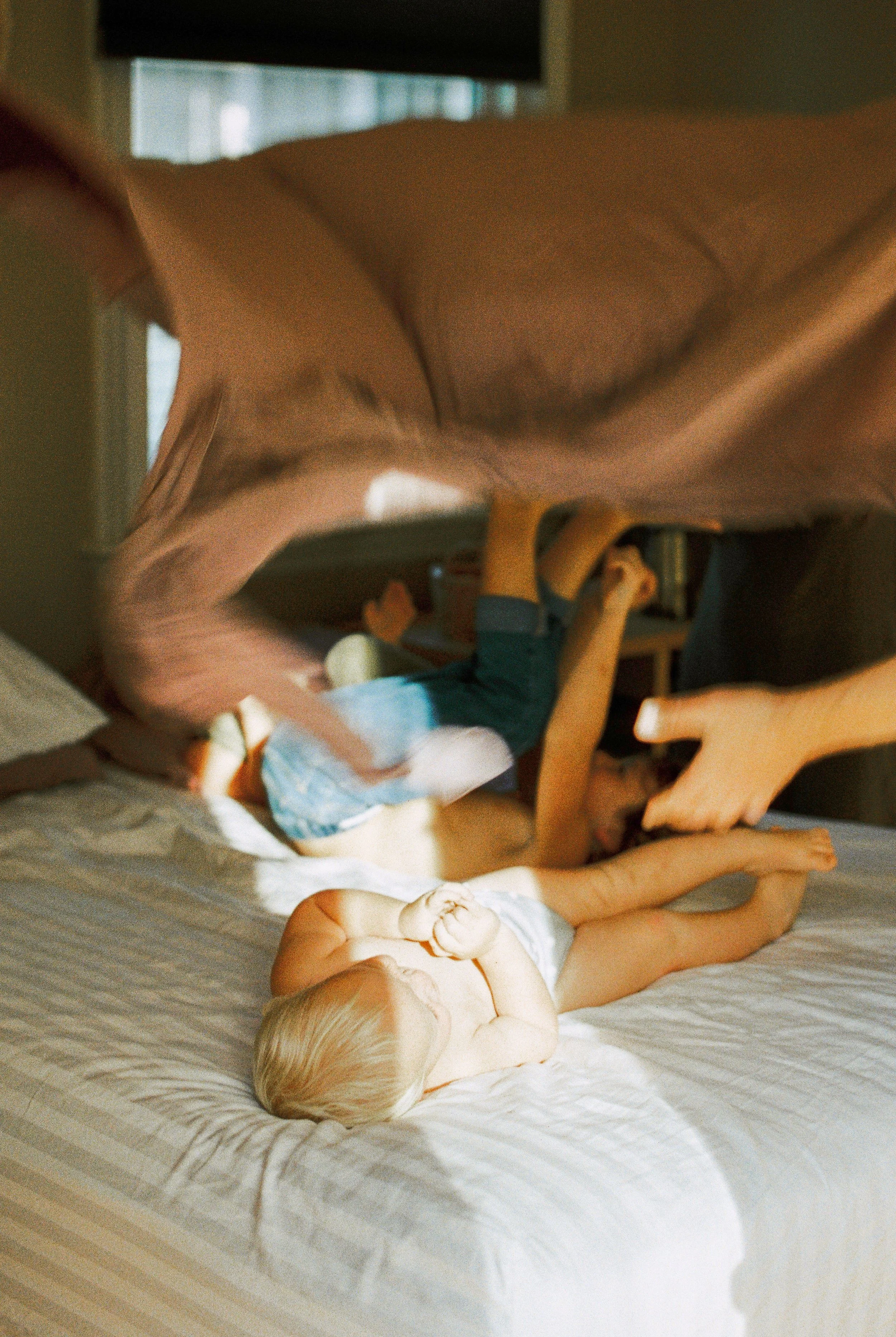 A baby with blonde hair lying on a bed, holding its foot, with a woman reaching towards the baby. The scene is indoors, with natural light coming through a window at a Bedford in hot family session. 