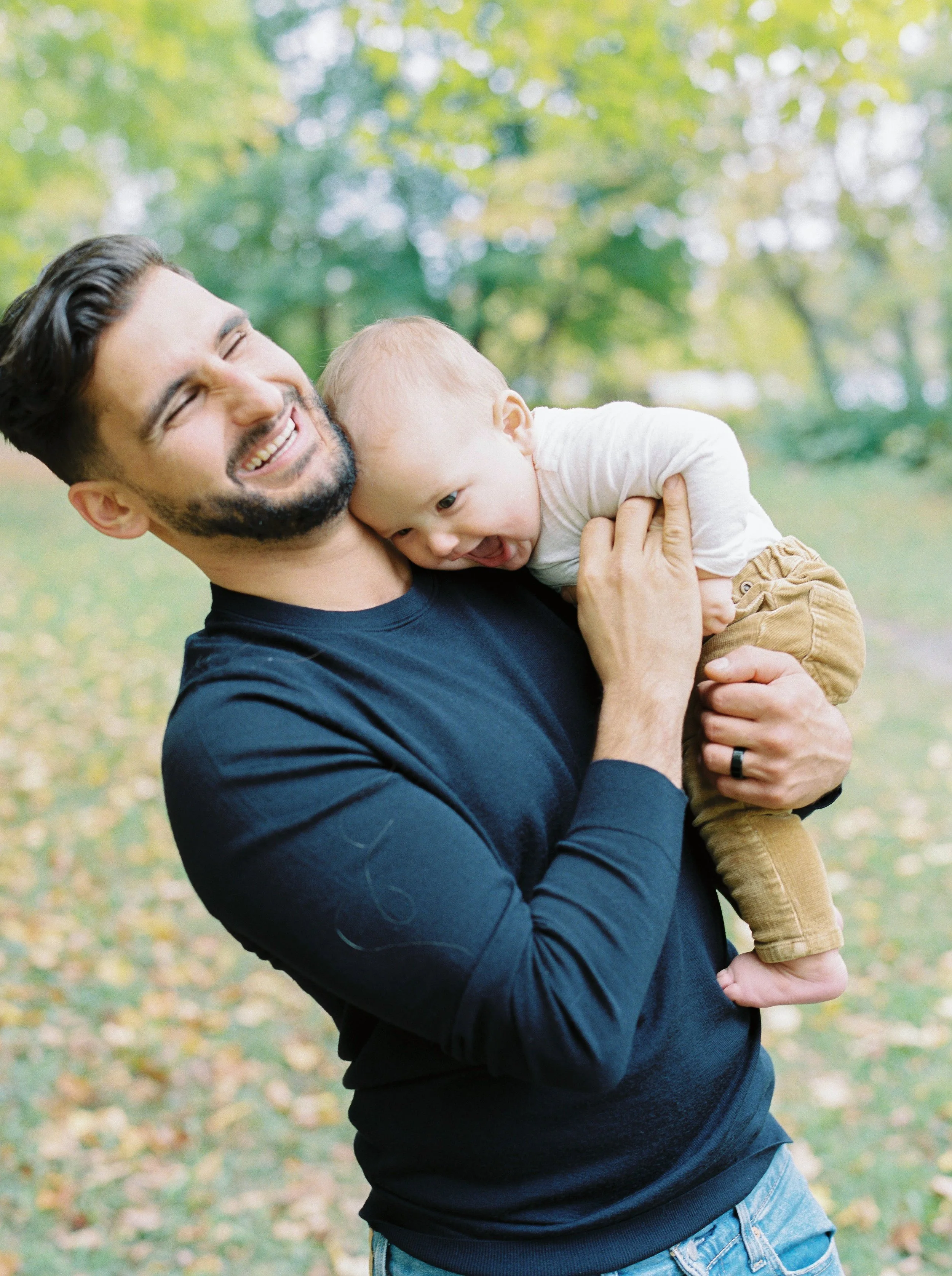 A man with dark hair and a beard smiling and holding a young child with blond hair, outdoors in a park with green and yellow trees in the background at a Dartmouth family photography session. 