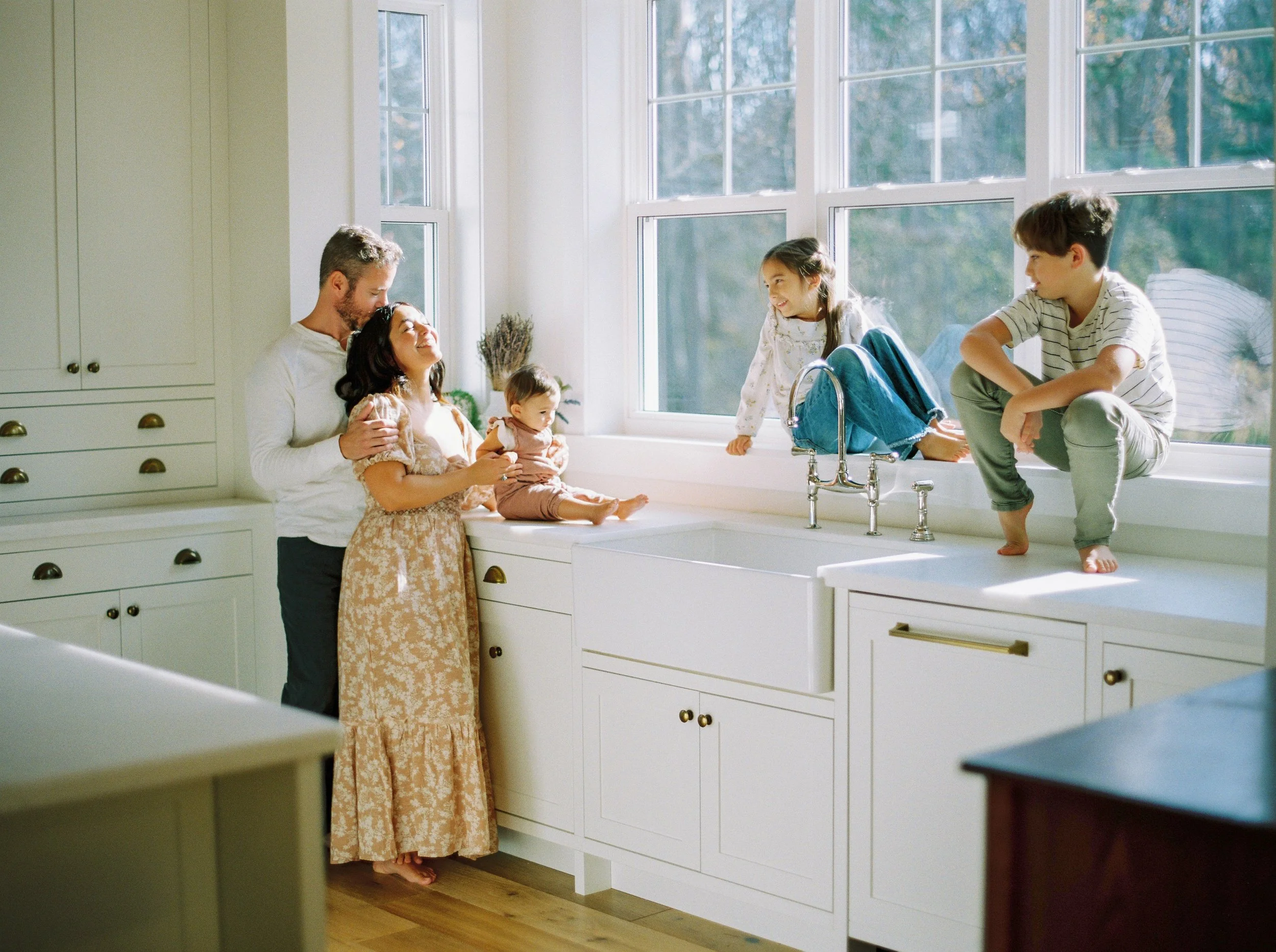 A family of five in a bright kitchen near a large window. The father and mother are sitting, with the mother holding their baby girl. Two older children are sitting on the kitchen counter, smiling and interacting at a Halifax in home family session.