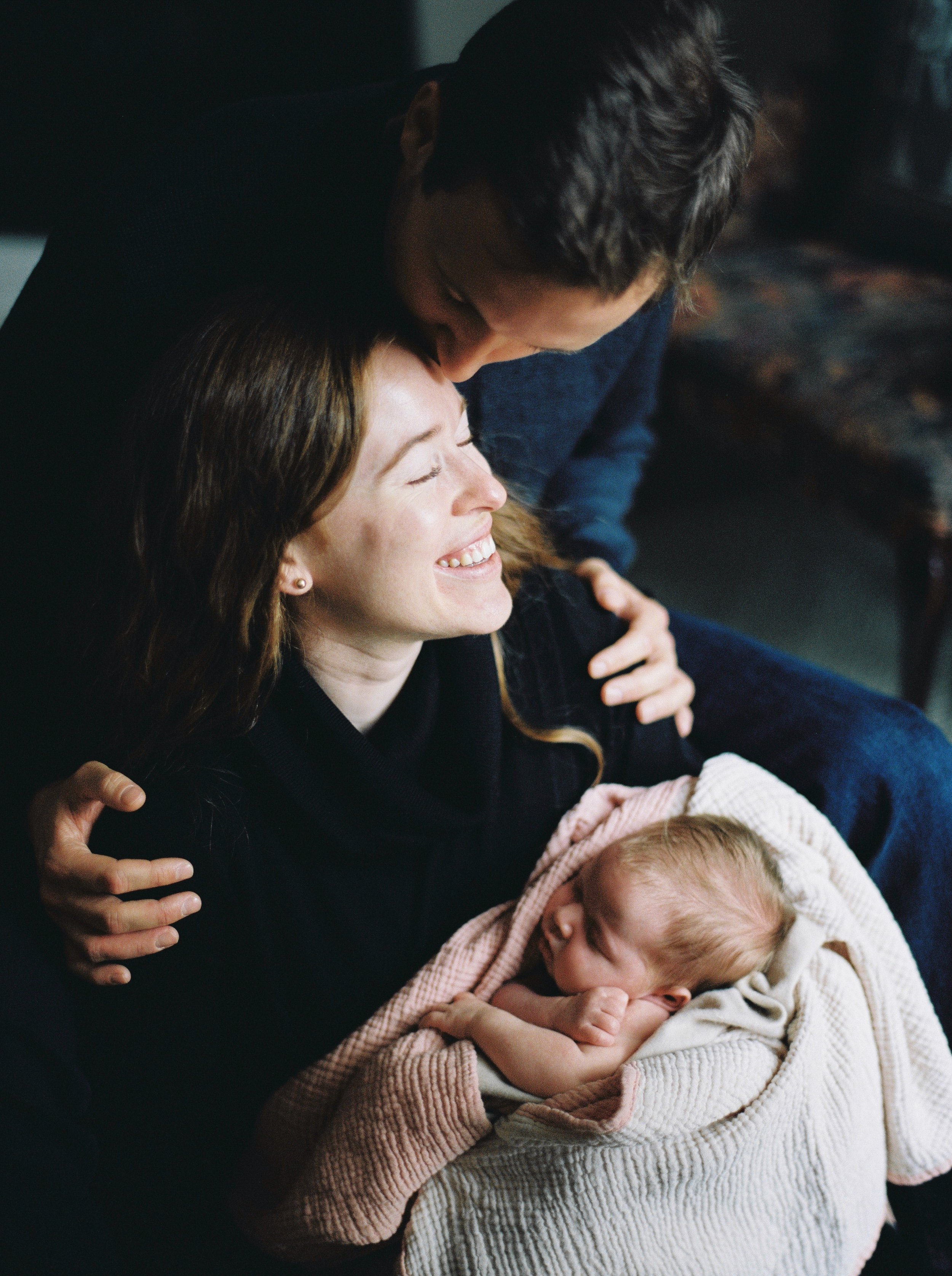 A woman with brown hair smiling and hugging a baby wrapped in a pink blanket, with an older person in the background in a fall river in home family photography session. 