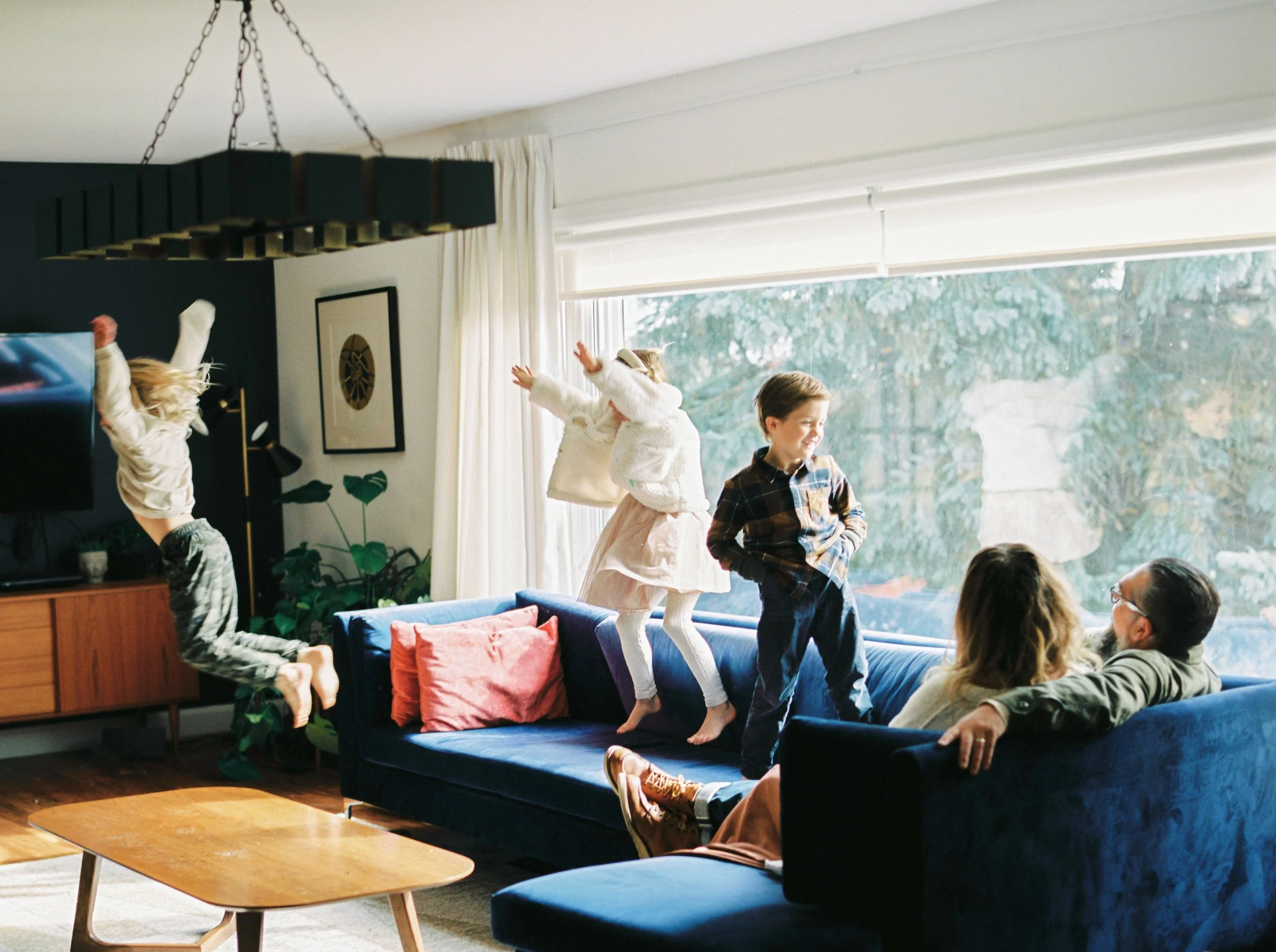 Children playing and jumping on a blue sofa inside a well-lit living room with large windows, curtains, and wall decorations at a Bedford in home family photography session