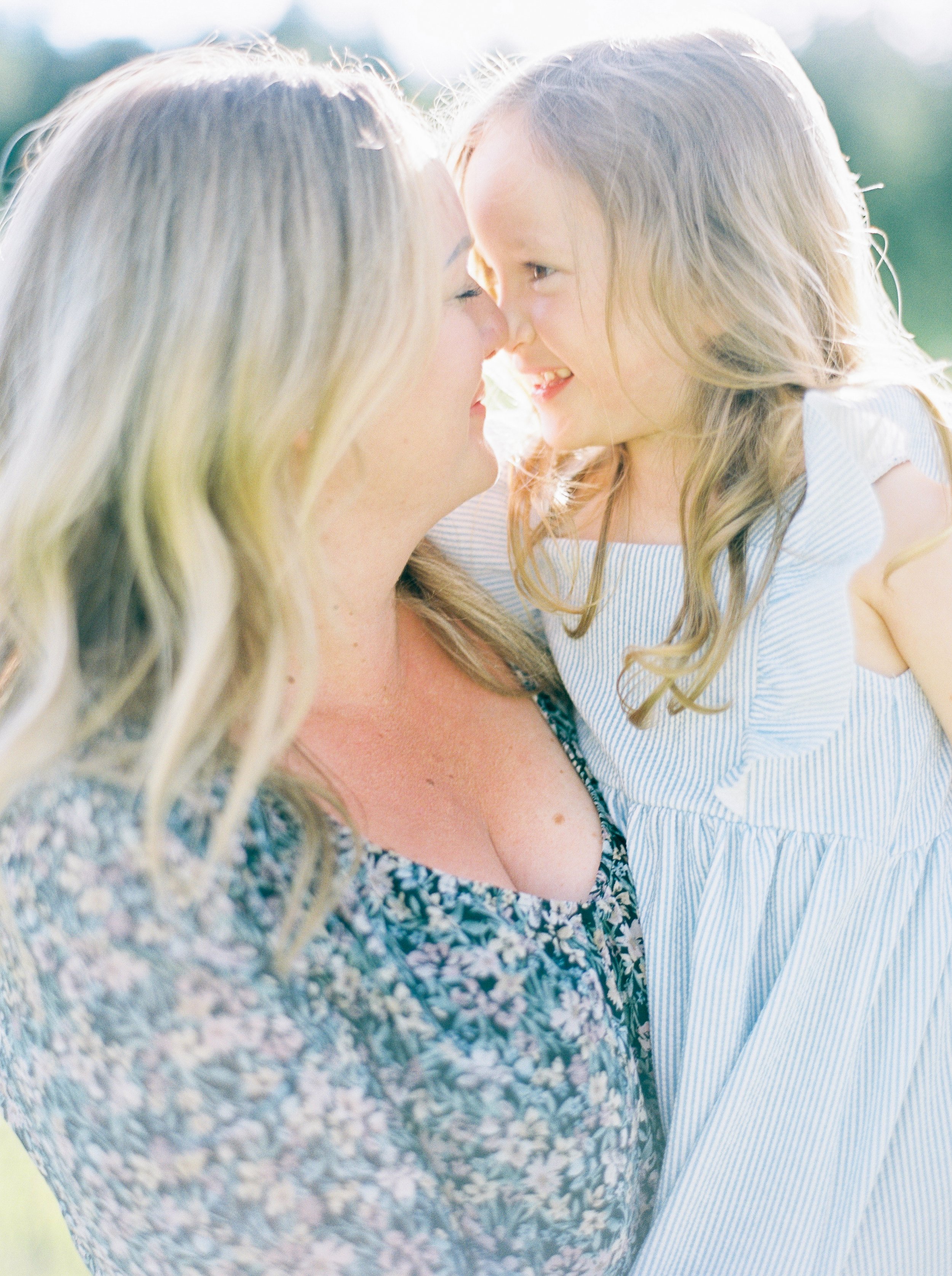 A close-up of a woman and a young girl with their foreheads touching and smiling, outdoors in natural light at a Bedford Family photography session. 