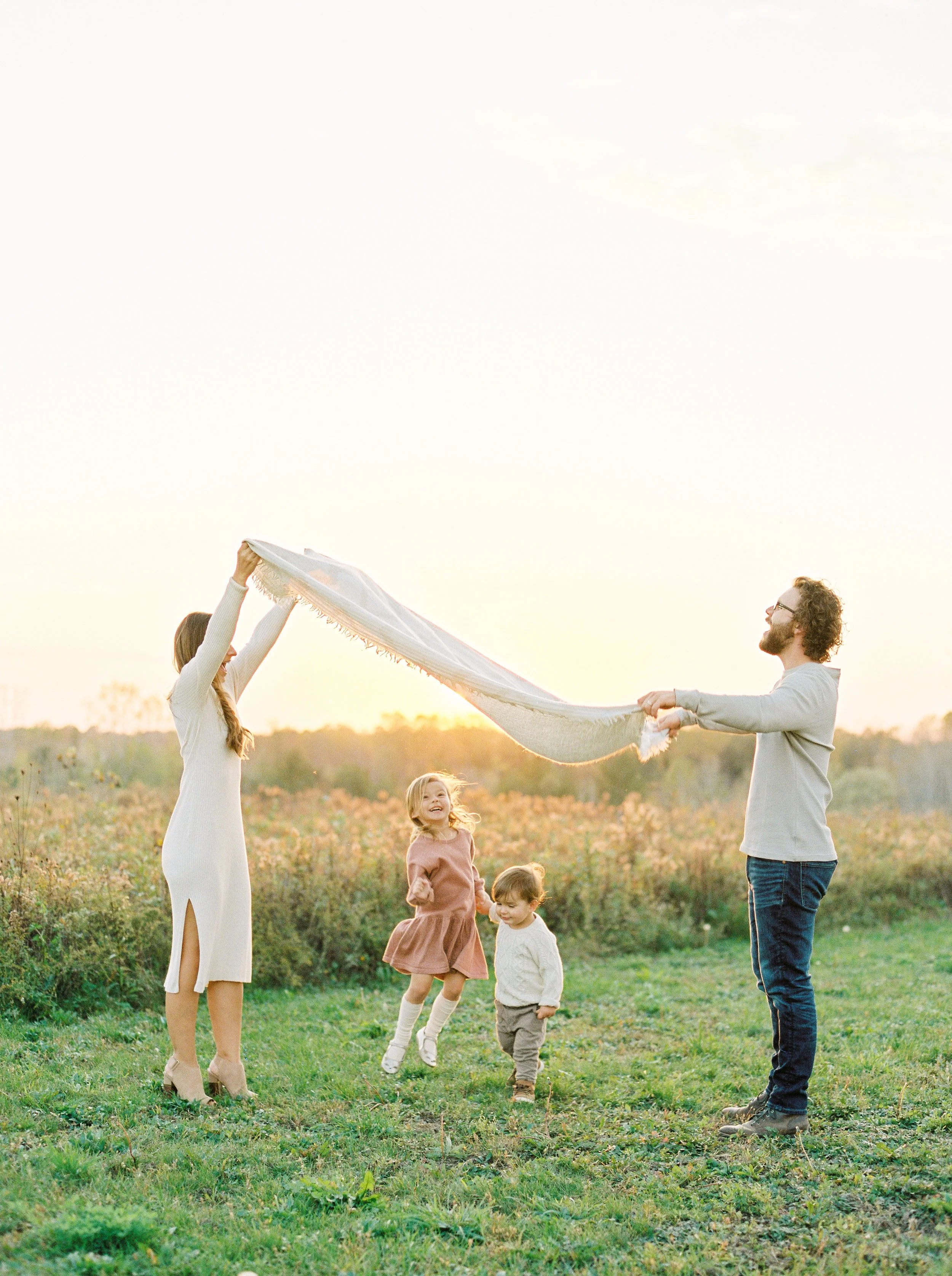 A family of five, including two young children, enjoying an outdoor moment during sunset in a grassy field. The woman on the left is holding a white cloth high, while the man on the right holds the other end. At a fall river NS family session.