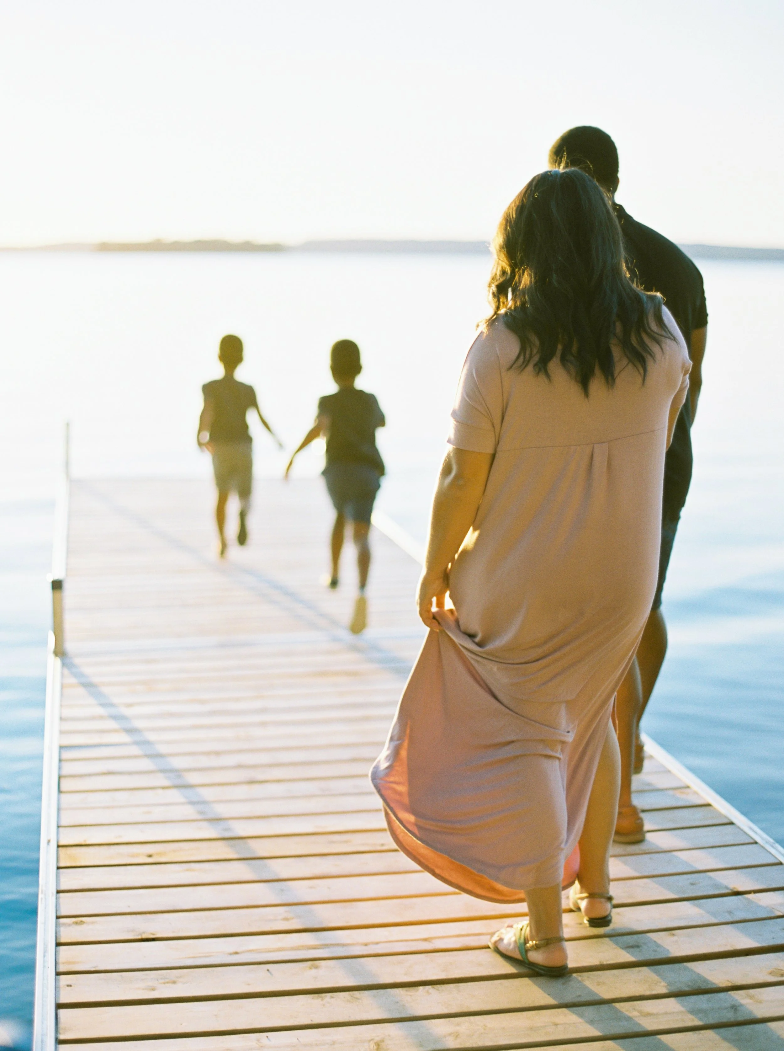 A woman in a pink dress and sandals walking on a wooden pier, with two children running in the background near a large body of water at sunset at a Lawrencetown family photography session. 