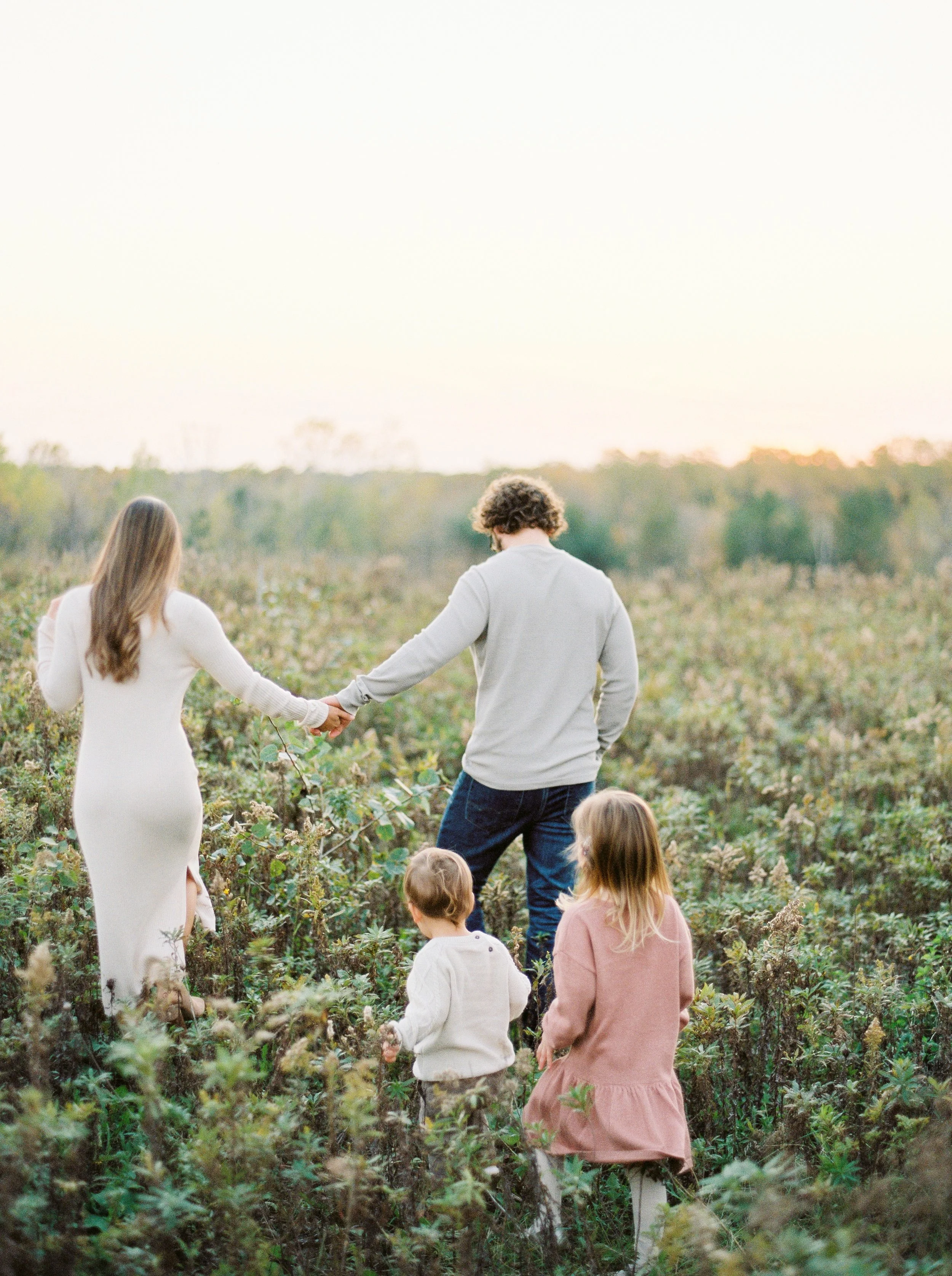 A woman and a man holding hands walking through a field of plants, with three children nearby, in an outdoor setting during sunset at a Halifax family photography session. 
