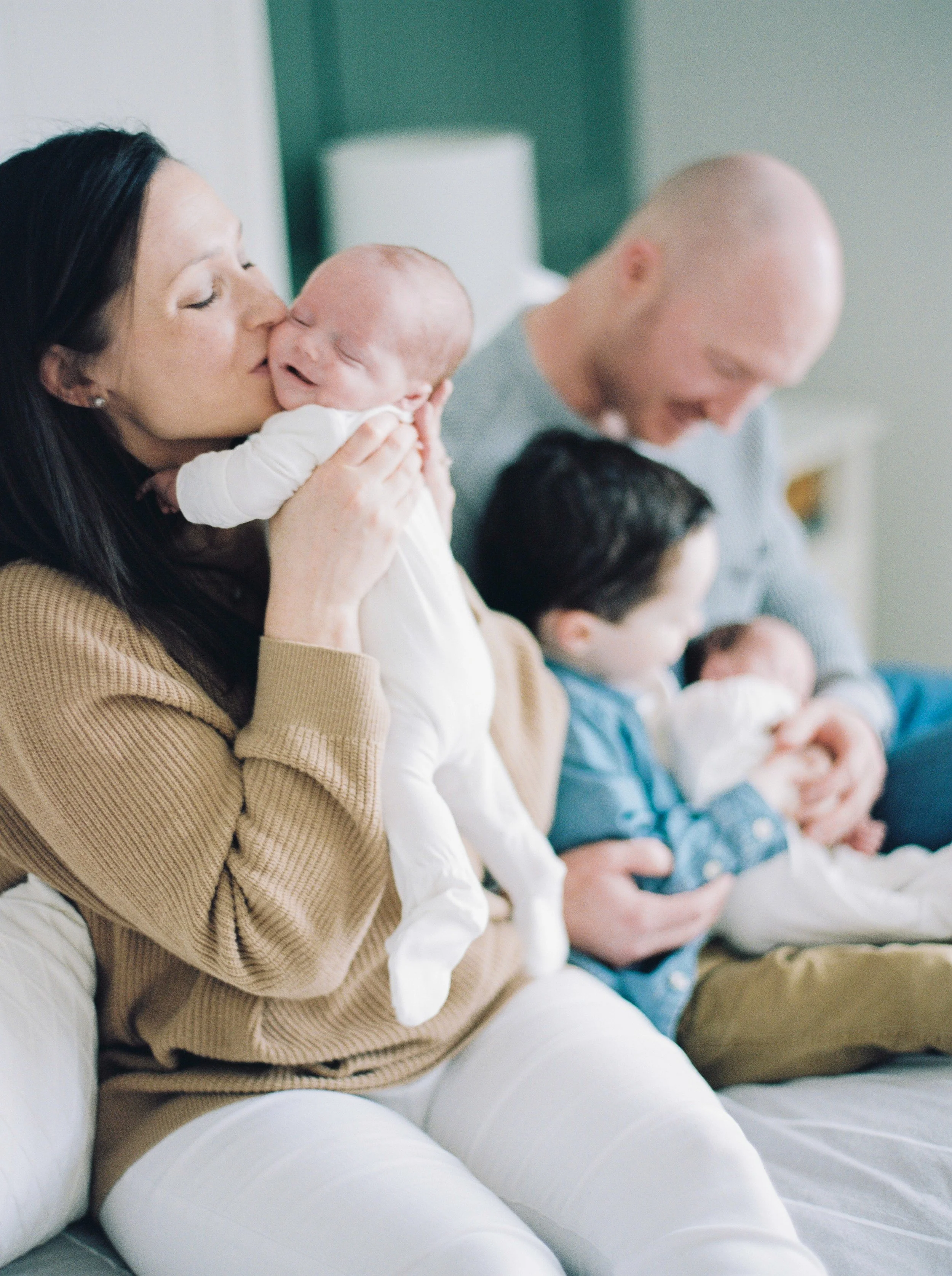 A woman with dark hair holding a newborn baby close to her face in a hospital room, next to a man with a tattoo on his arm holding another baby, all sitting on a white couch at a Bedford family photography session. 