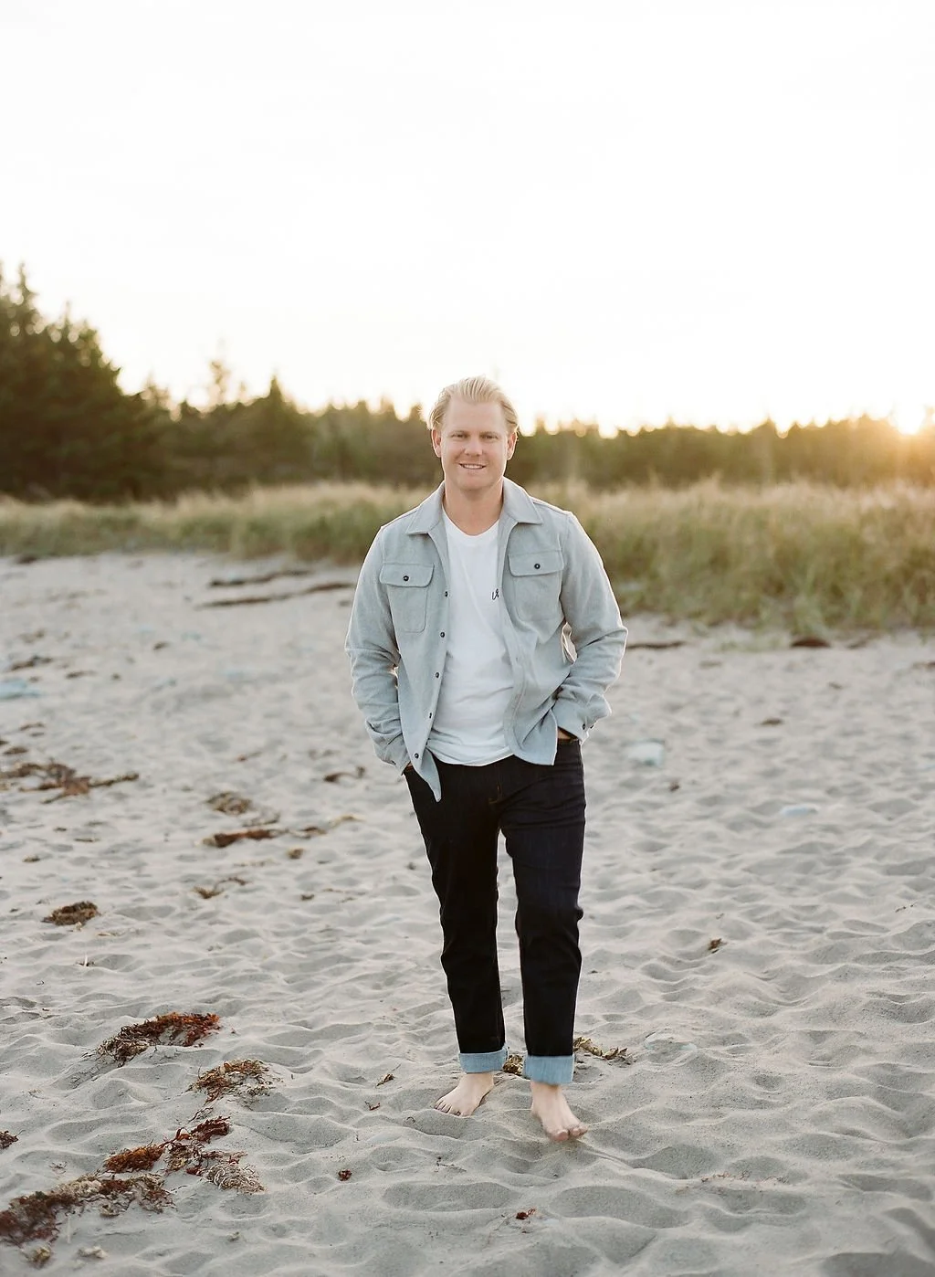 A young man with blonde hair walking barefoot on a sandy beach during sunset, wearing a light gray jacket over a white t-shirt and rolled-up black jeans.