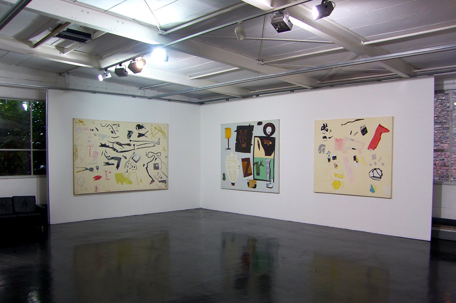 An art gallery with white walls displaying three abstract paintings, a black seating area in the corner, and windows showing trees outside. The gallery has a polished dark floor and professional ceiling lighting.
