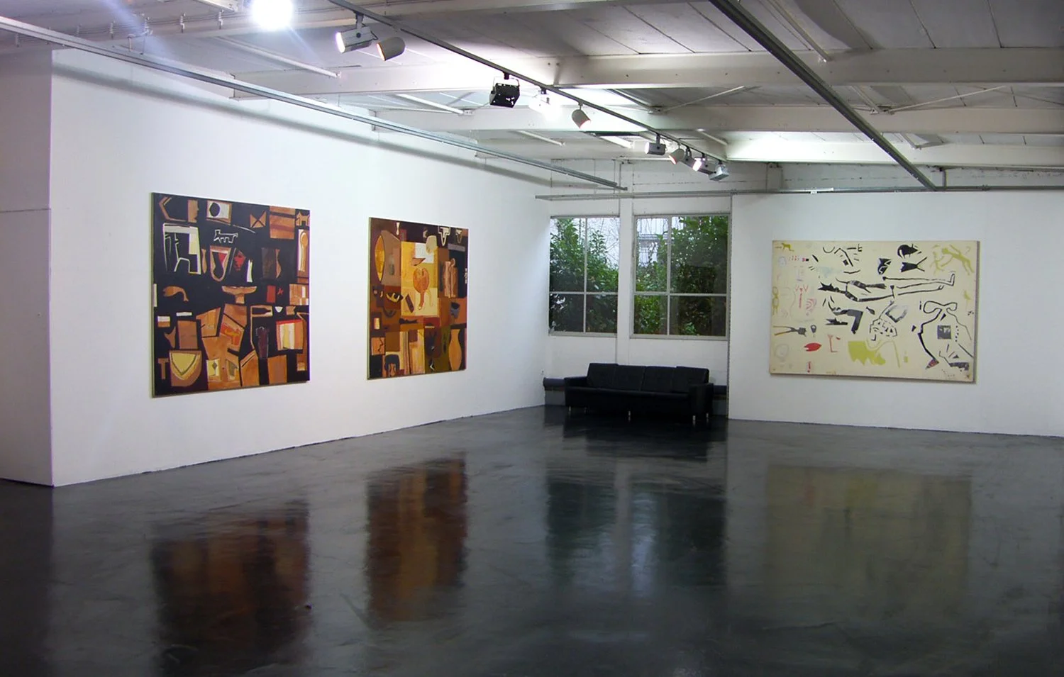 An art gallery with three abstract paintings on white walls, hardwood floors, and a black sofa near the corner beneath two windows showing outside greenery.