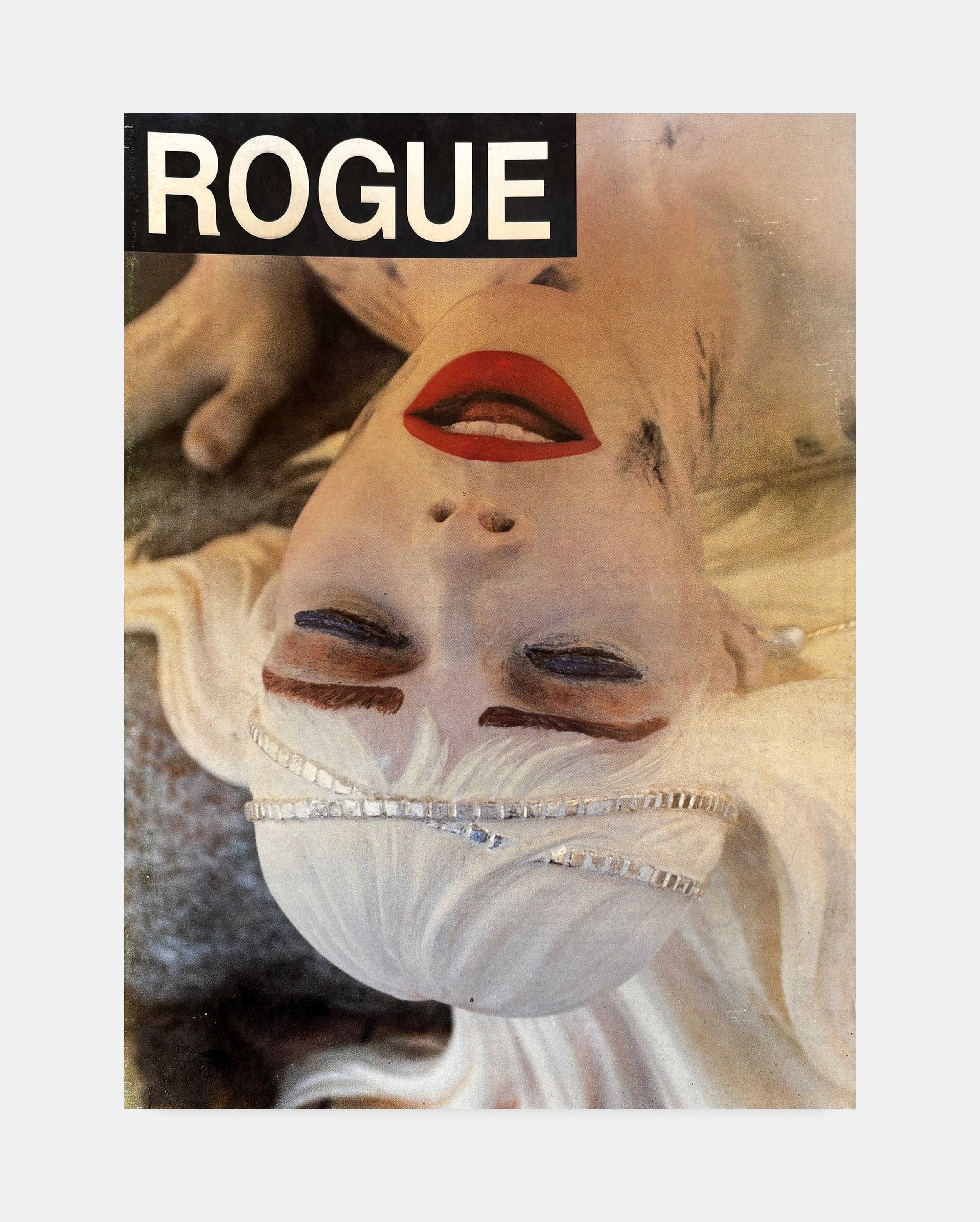 Upside-down image of a woman with red lipstick, dark eye makeup, with blonde hair, wearing a white top with silver embellishments, lying down on a textured surface, with the word 'ROGUE' in white letters on a black rectangle at the top.