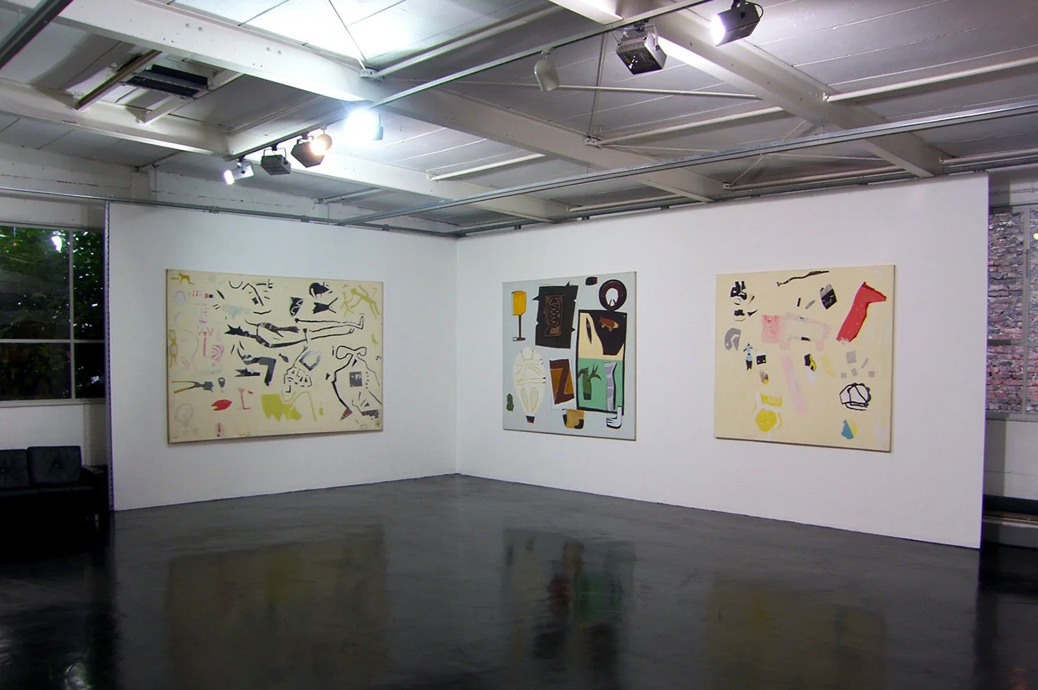 An art gallery with three abstract paintings hanging on a white wall, with black floor and ceiling lights, and a window showing trees outside.