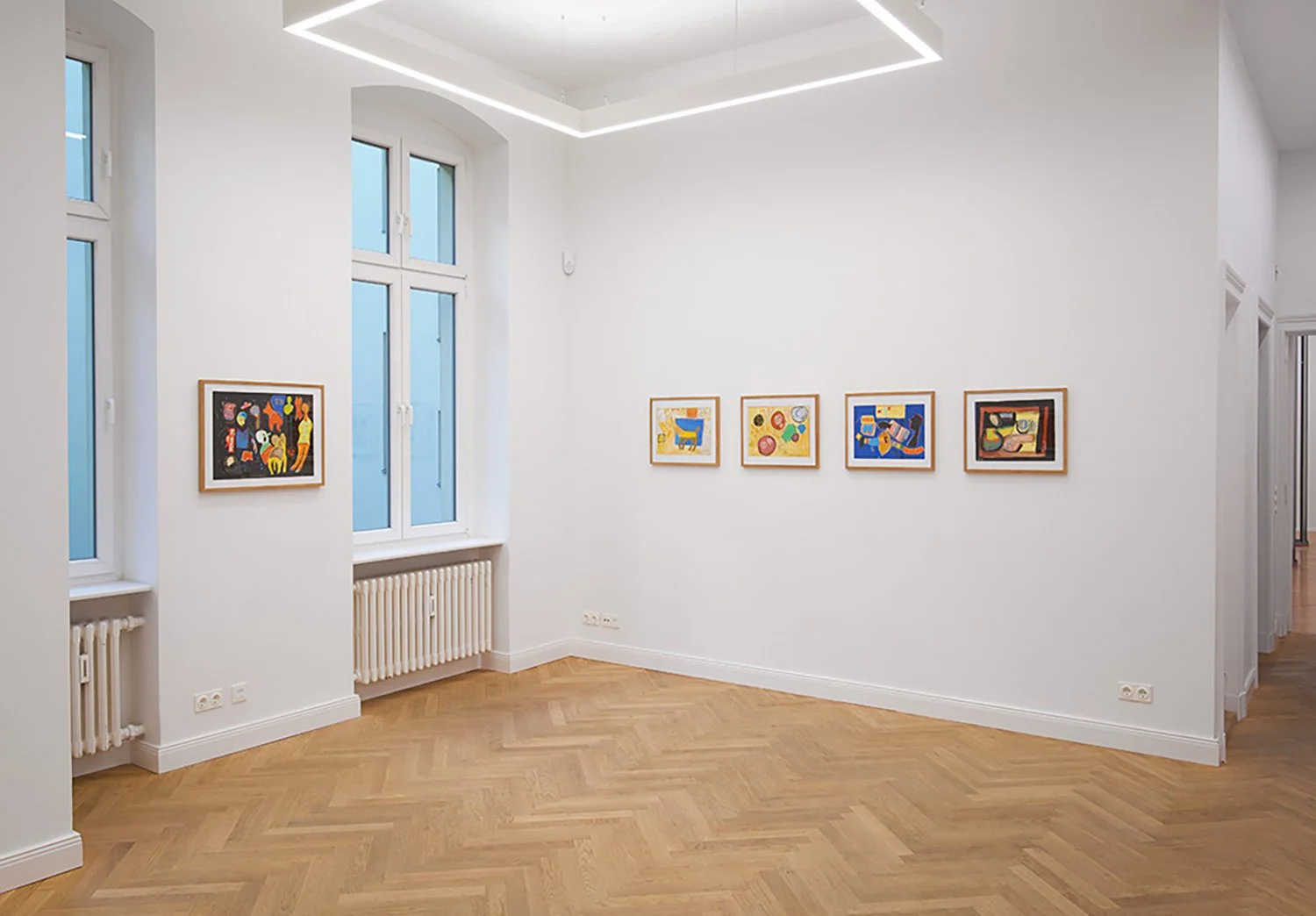 An art gallery with a white wall decorated with five colorful paintings. The room has large windows with white frames, wooden herringbone flooring, and minimal lighting.