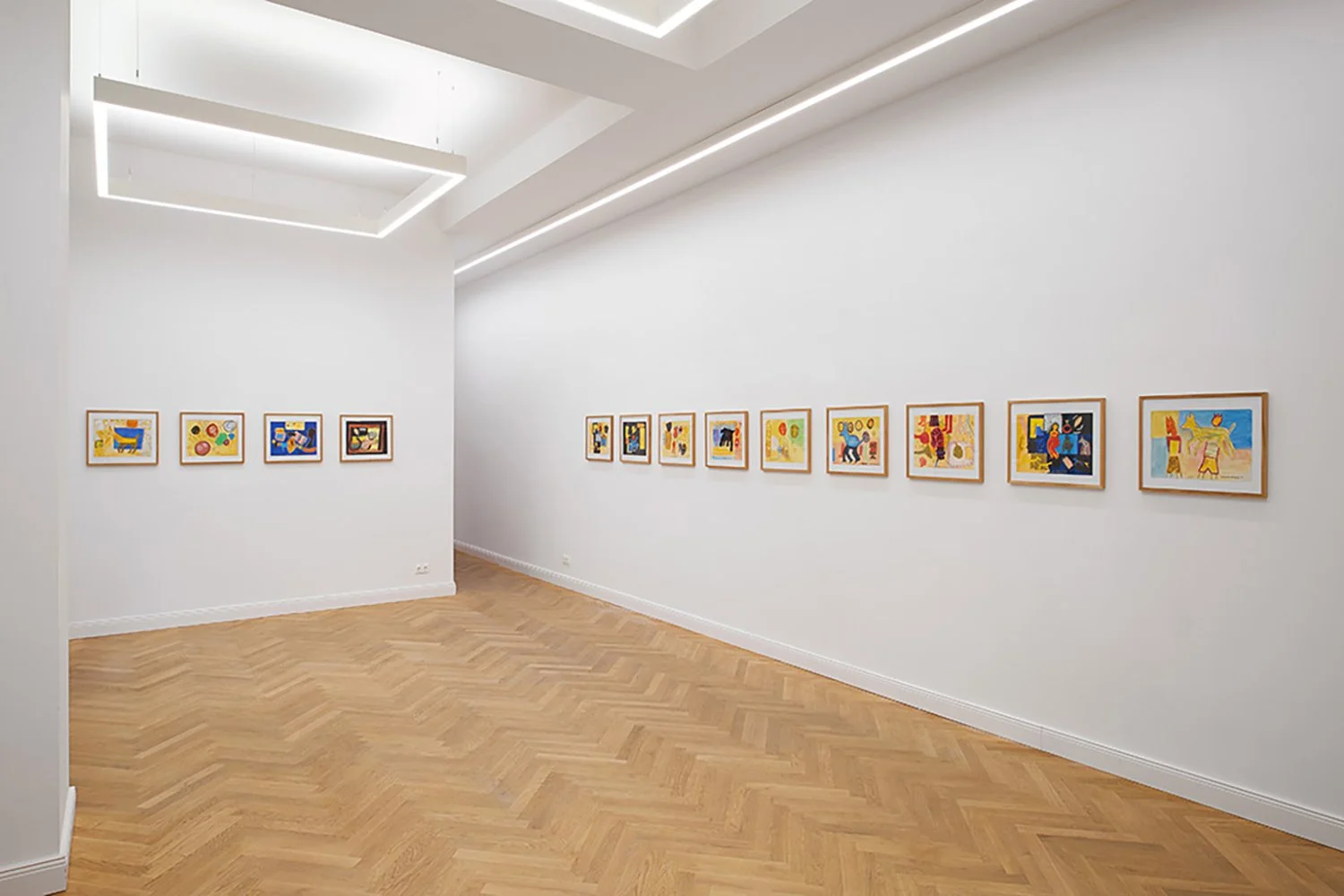 Art gallery with colorful framed paintings displayed in a well-lit white room and wooden parquet flooring.