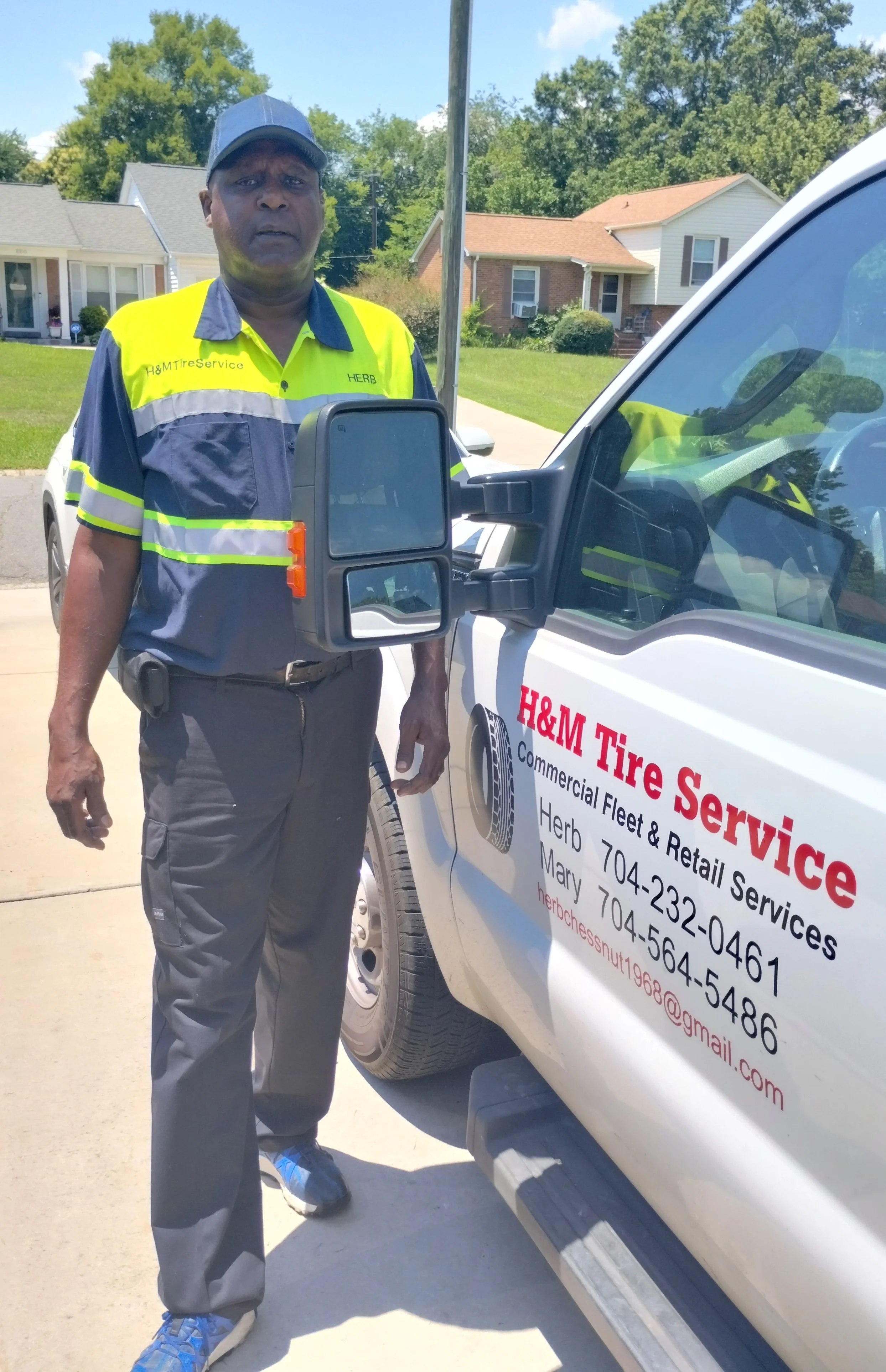 A man wearing a yellow and gray reflective safety uniform standing next to a white service truck with red lettering. The man is outdoors on a sunny day, with houses, trees, and a clear blue sky in the background.